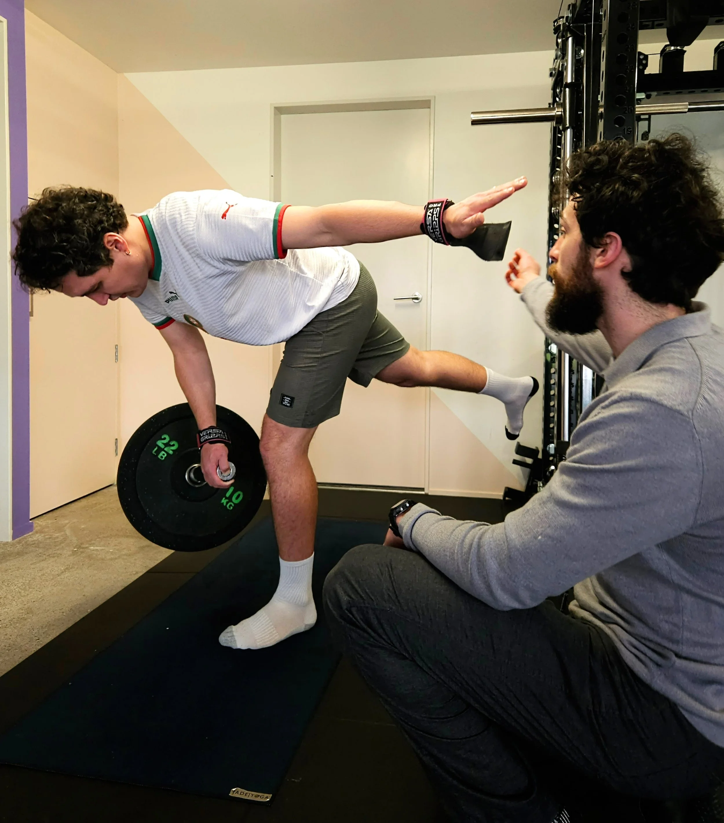 A man in athletic clothing doing a landmine singe-leg romanian deadlift. Trained by Julian Etchegoin, Craft Fitness Studio Founder and personal trainer. The gym has purple walls and various workout equipment.