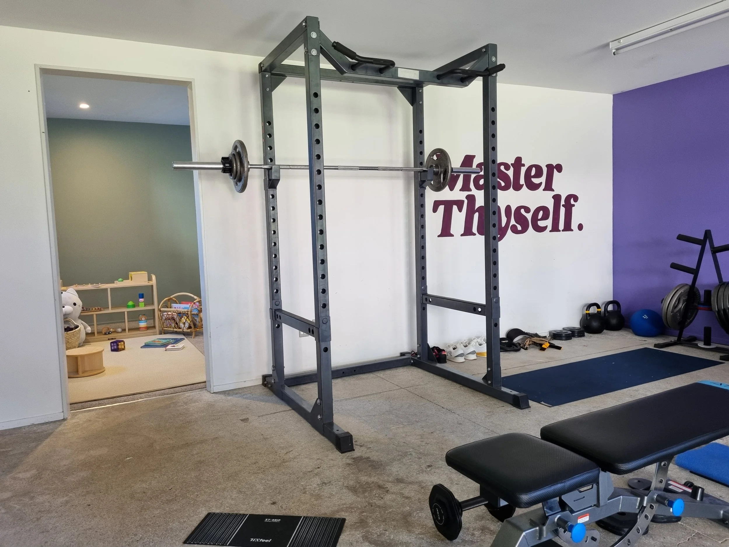 Fitness room with squat rack, barbells, kettlebells, medicine balls, and workout benches, adjacent to a children's play area with stuffed animals and toys.