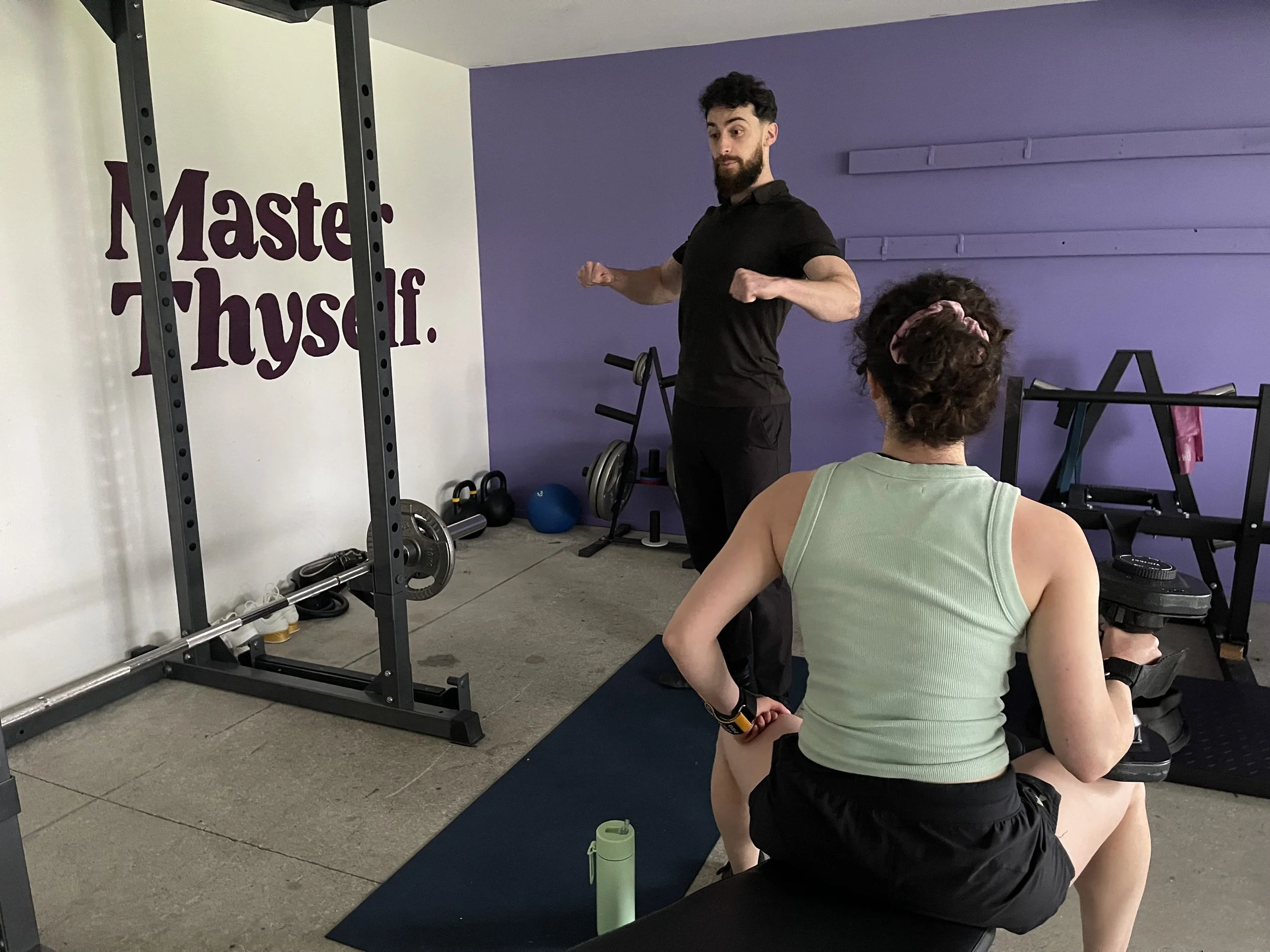 A woman sitting on a workout bench holding a dumbbell in a gym, watching a man demonstrating an exercise in front of a wall with motivational text. The wall has a reflection of gym equipment, and the text says, 'Master Yourself.'