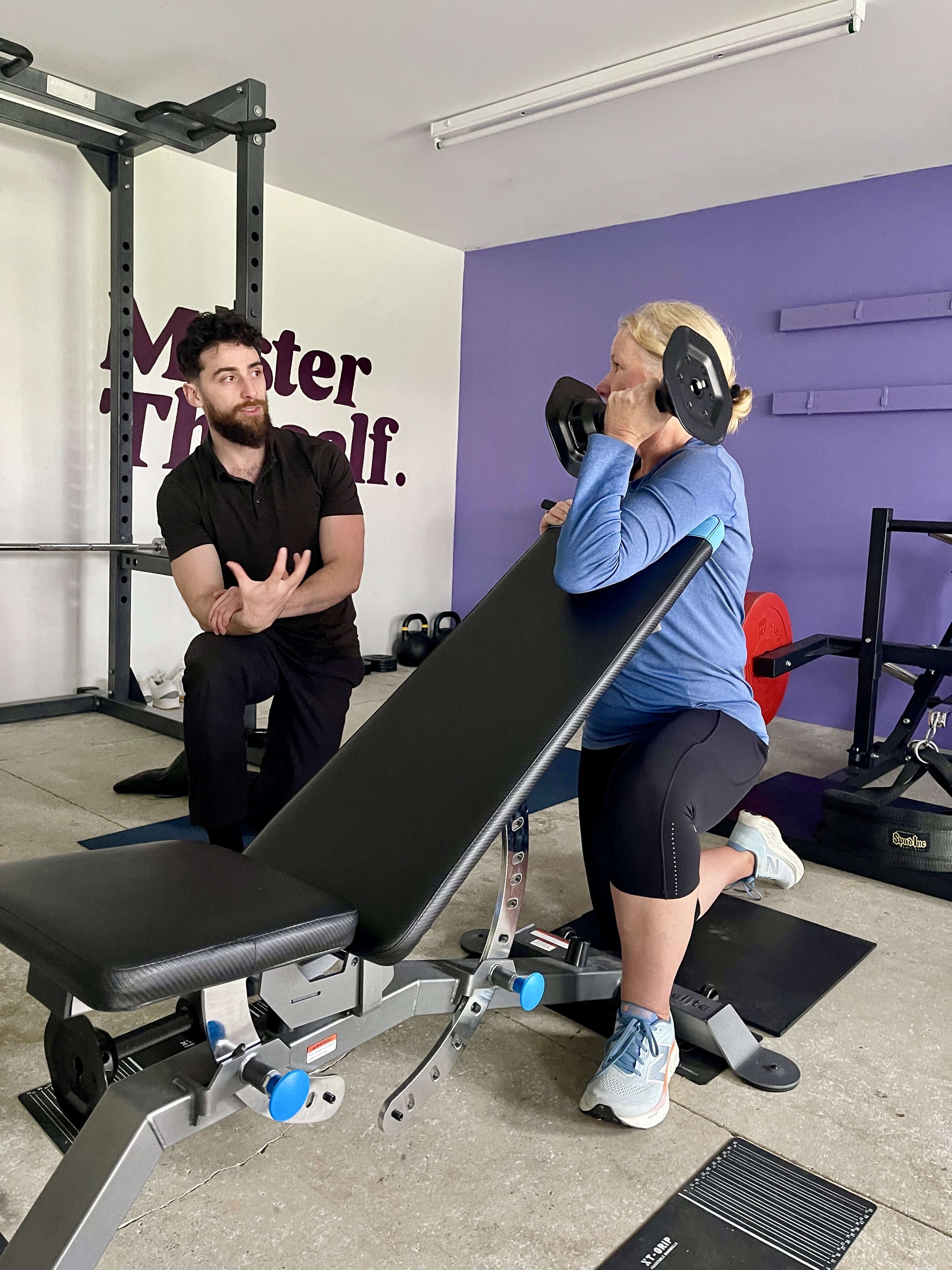A woman performing a kneeling weightlifting exercise with a dumbbell on her shoulder, while a trainer observes and gives instructions in a gym with purple and white walls.