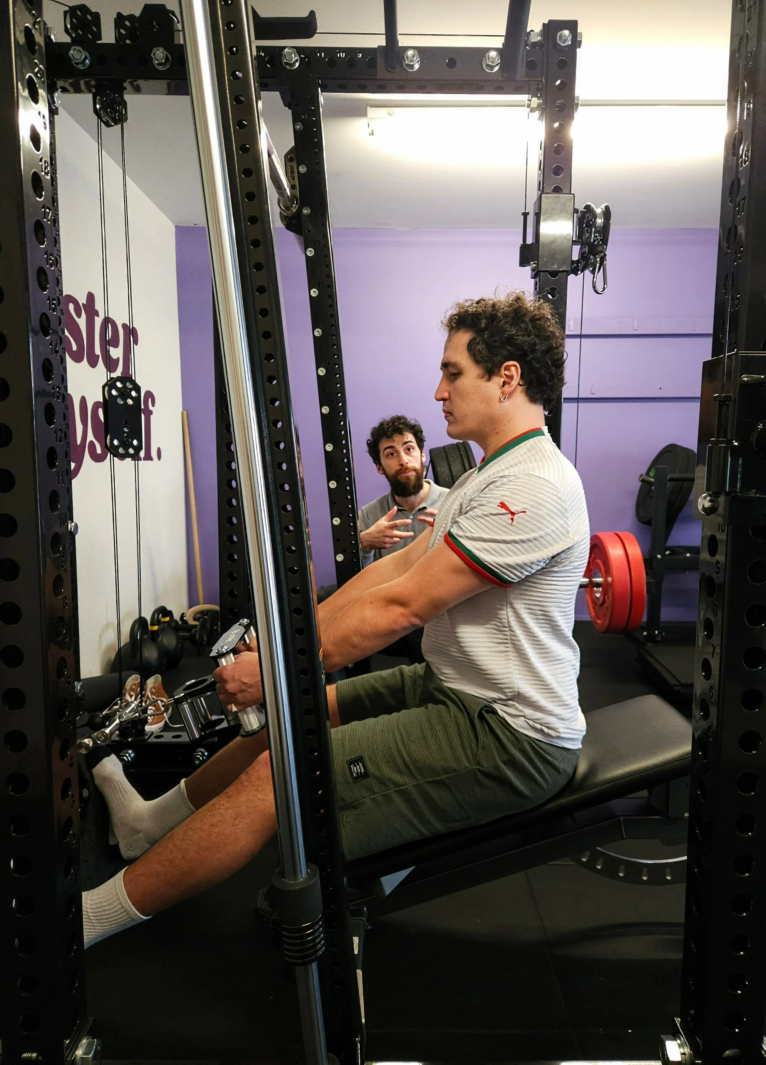 A man in athletic clothing preparing to do a seated cable row with the REP Ares™ 2.0 cable attachment. Trained by Julian Etchegoin, Craft Fitness Studio Founder and personal trainer. The gym has purple walls and various workout equipment.