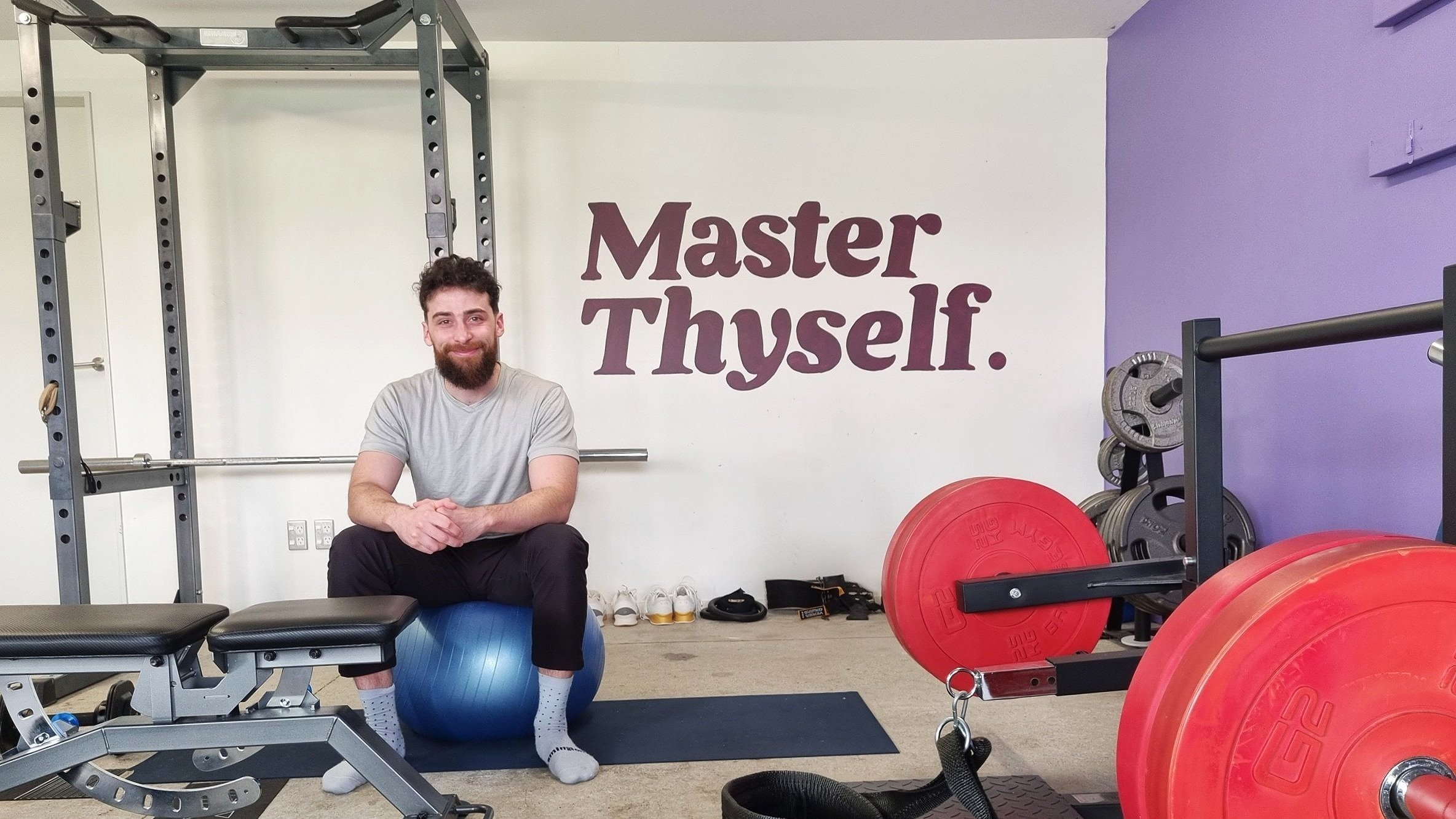 A man with a beard and gray t-shirt sitting on a blue exercise ball in Craft Fitness | Personal Training Wellington gym, smiling at the camera with the words 'Master Thyself.' on the wall behind him.