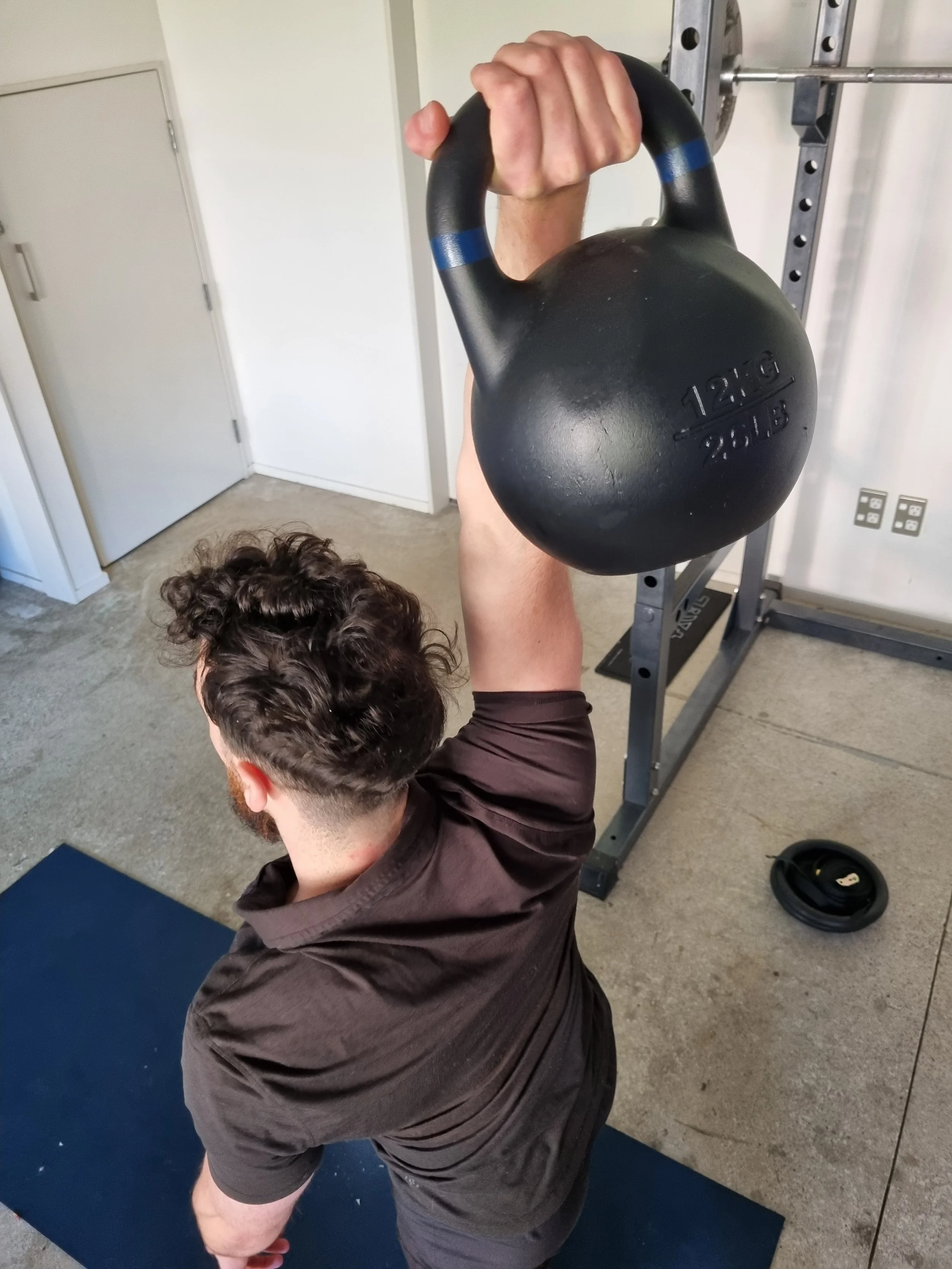 A man in a black shirt is lifting a heavy black kettlebell labeled 12 kg / 26 lb while on a yoga mat in a gym. The man is on one knee, and a weight rack and door are visible in the background.
