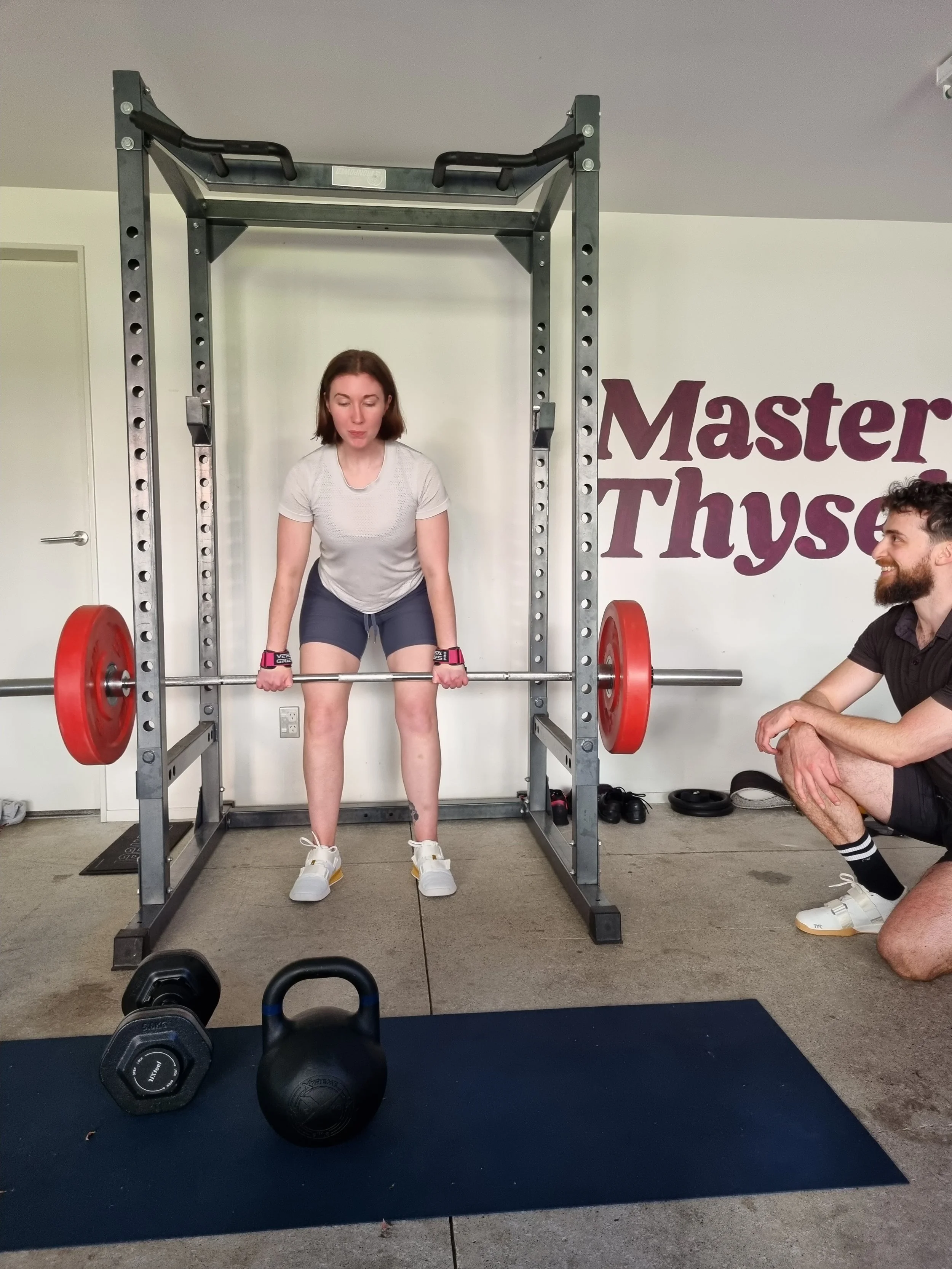 A woman performs a deadlift with a barbell and red weight plates in a gym, monitored by a trainer squatting nearby, with kettlebells and dumbbells on the floor.