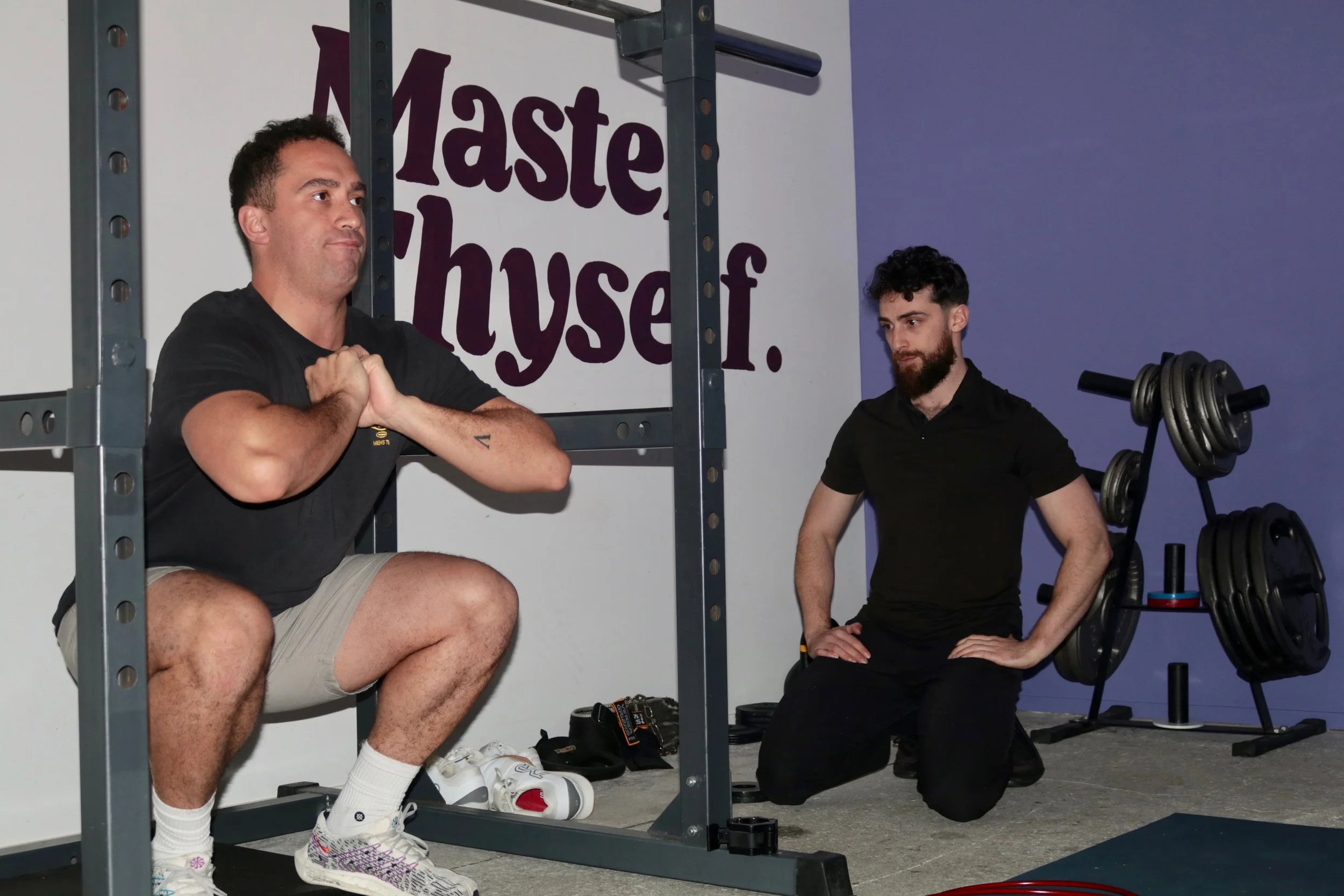 A man in workout attire squat exercises while a trainer observes him in a gym with a sign that reads 'Master Thyself' in the background.