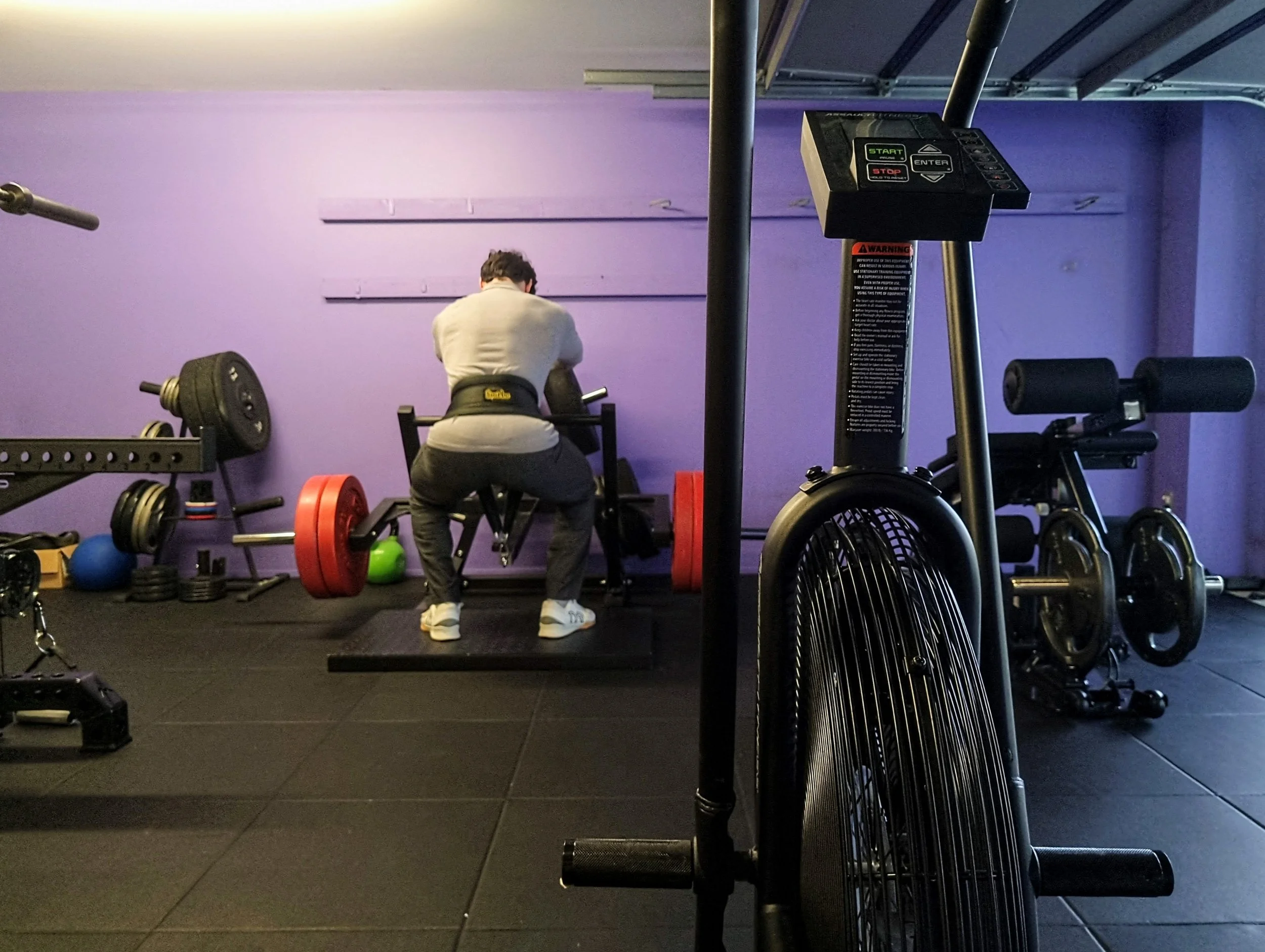 Julian Etchegoin, founder and personal trainer at Craft Fitness Studio, using the belt squat machine in his Karori Studio. Air bike and other gym equipment in the background, purple branding.