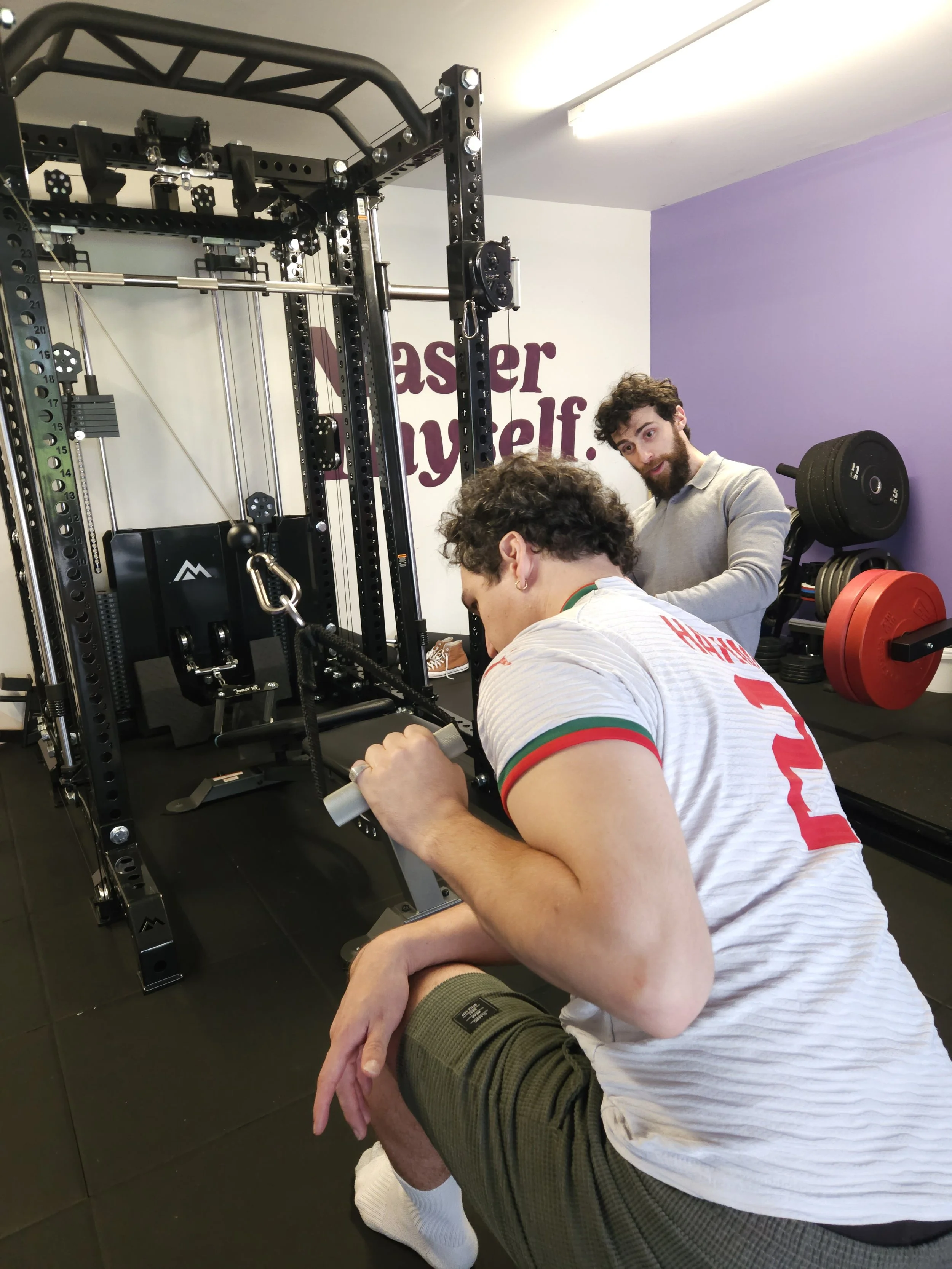 A man in athletic clothing doing a single arm lateral pulldown with the REP Ares™ 2.0 cable attachment. Trained by Julian Etchegoin, Craft Fitness Studio Founder and personal trainer. The gym has purple walls and various workout equipment.