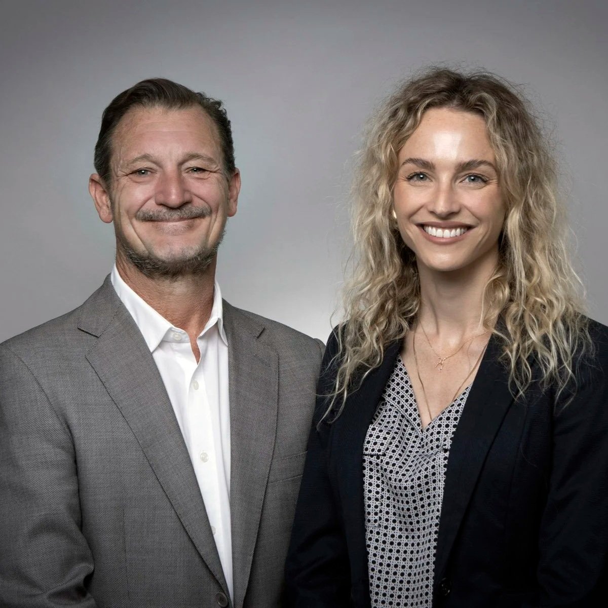 A man and woman standing side by side, smiling at the camera, in a professional setting with a plain gray background.