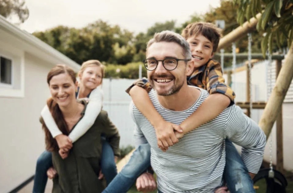 A smiling family of four outdoors, with father giving piggyback rides to his two children, while the mother walks beside them, all appearing happy and enjoying a sunny day.