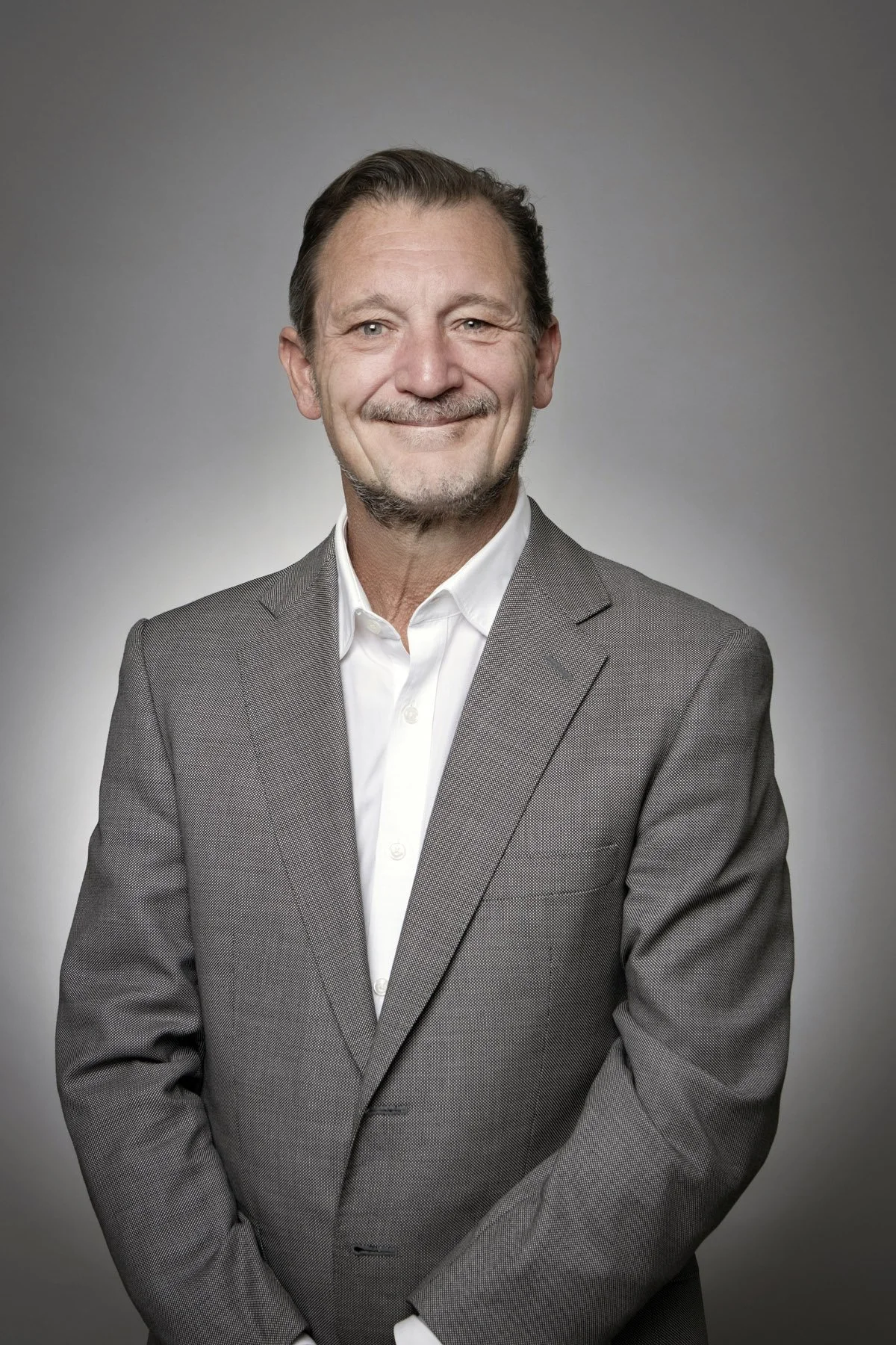 A portrait of a smiling middle-aged man with brown hair and a beard, wearing a gray suit jacket and white shirt, standing against a gray background.