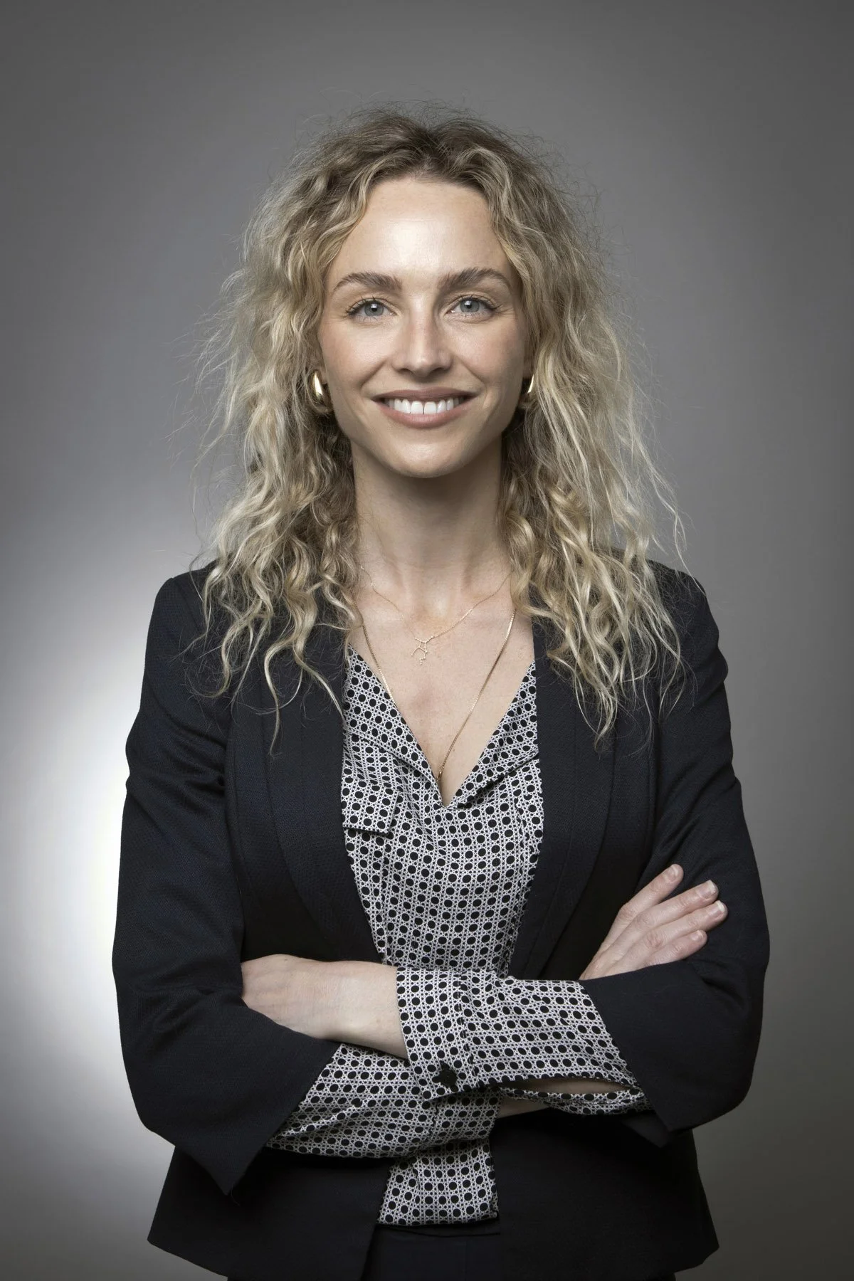 A young woman with curly blonde hair, wearing a black blazer over a patterned blouse, standing with arms crossed and smiling at the camera with a gray background.