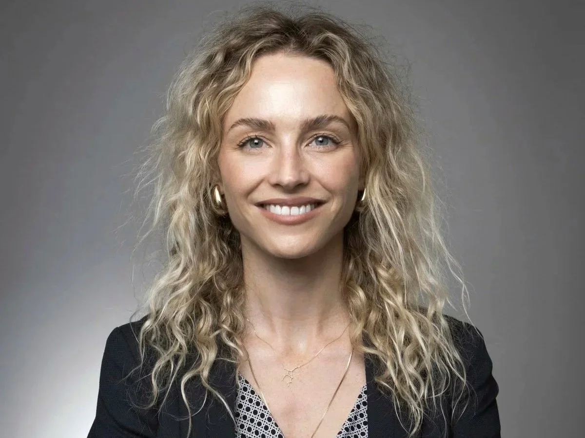 A woman with curly blonde hair, blue eyes, and light skin, smiling at camera, wearing a black blazer, white patterned top, gold hoop earrings, and two necklaces, against a plain gray background.