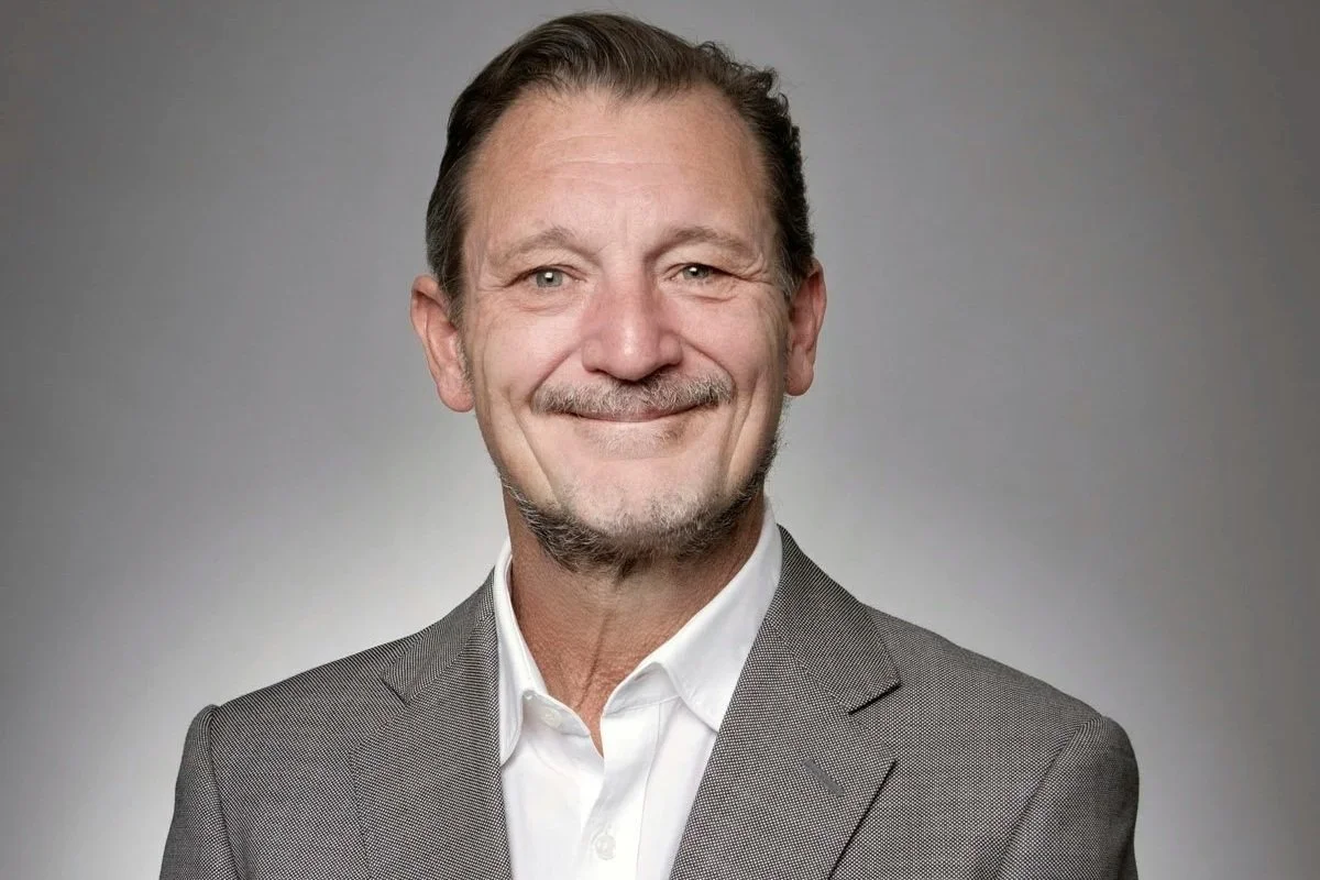 A middle-aged man with light brown hair, a mustache, and a goatee, wearing a gray suit and white shirt, smiling against a neutral gray background.