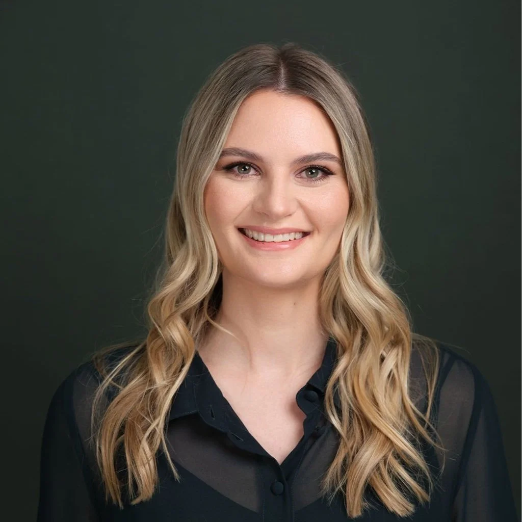 A woman with long wavy blonde hair smiling, wearing a black blouse with sheer sleeves, against a dark green background.