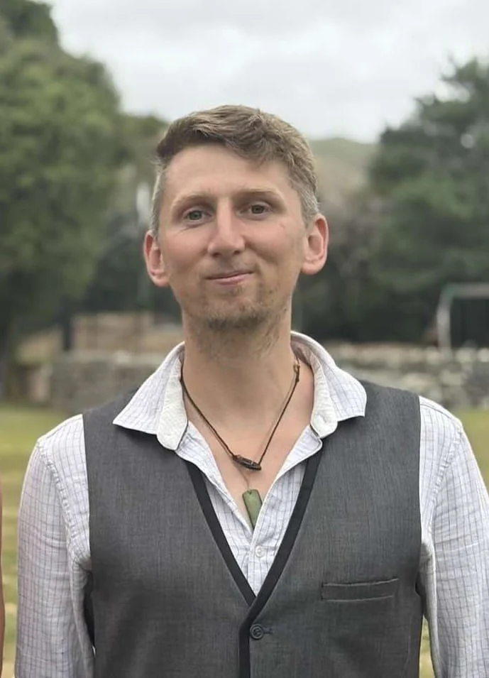 A young man with light brown hair, wearing a white collared shirt under a gray vest, stands outdoors with trees and a cloudy sky in the background. He has a necklace with a green pendant.