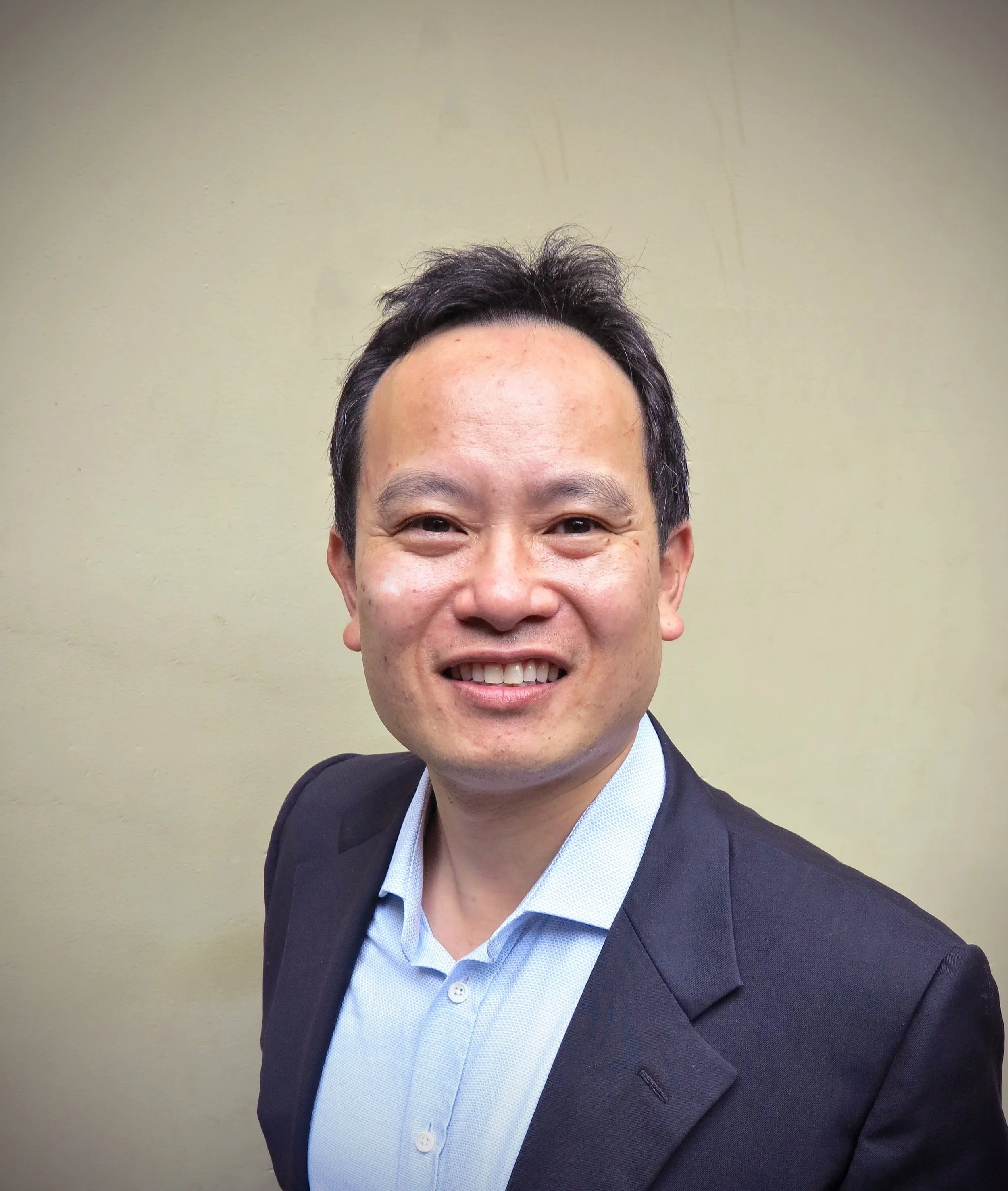 Headshot of a smiling man in a suit and light blue shirt against a plain wall.