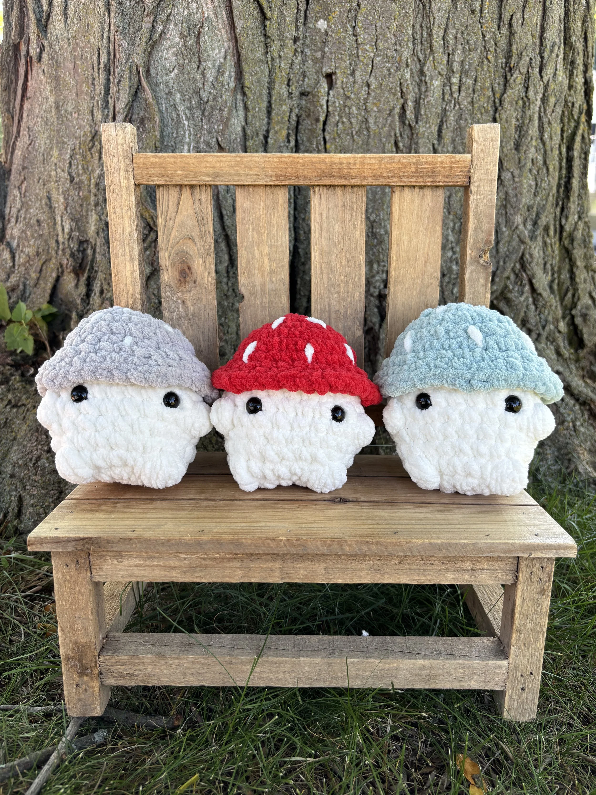 Three crochet mushroom plushies in red, grey, and blue on wooden bench