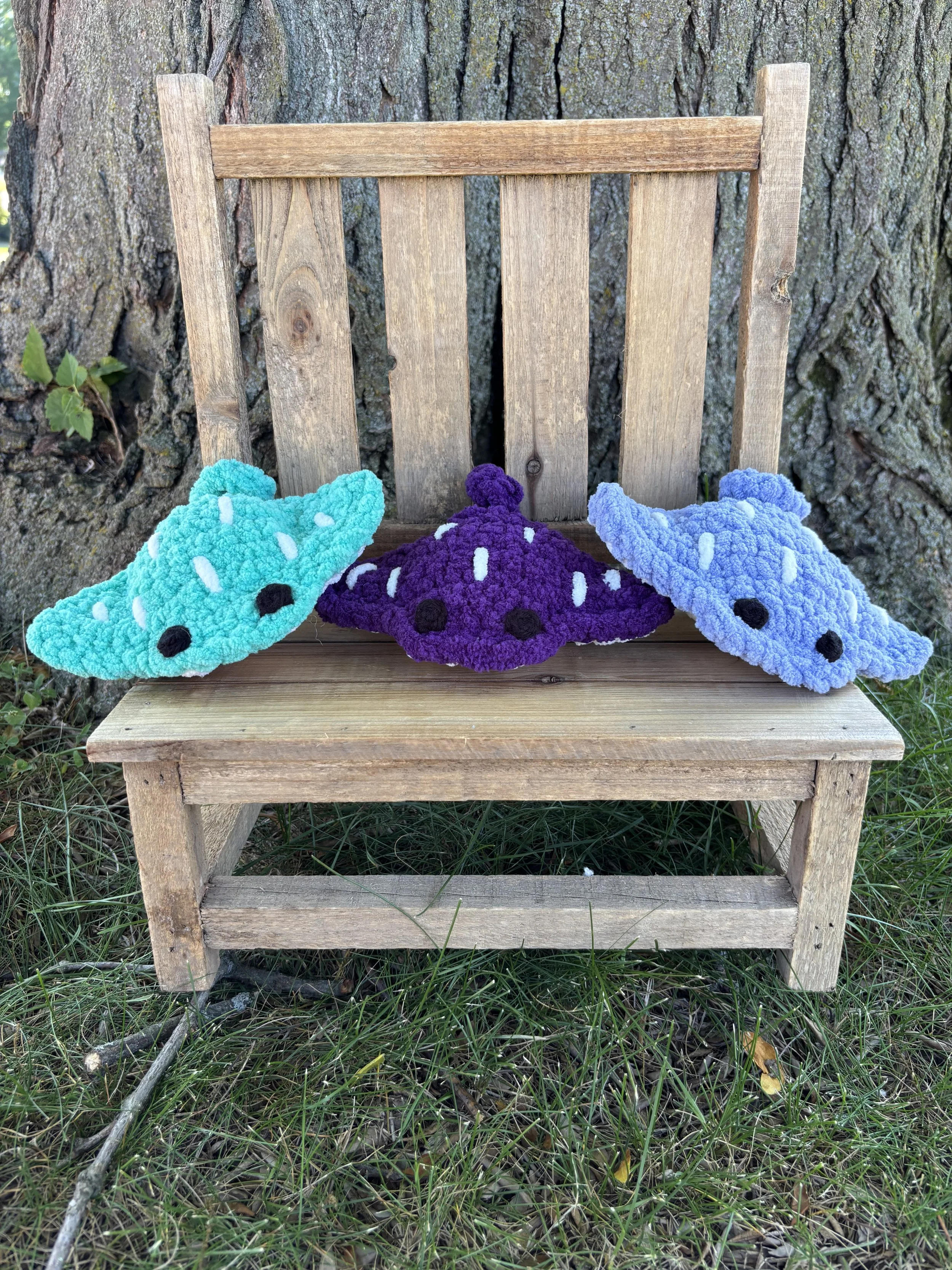 Three crochet stingray plushies in teal, purple, and blue on wooden bench