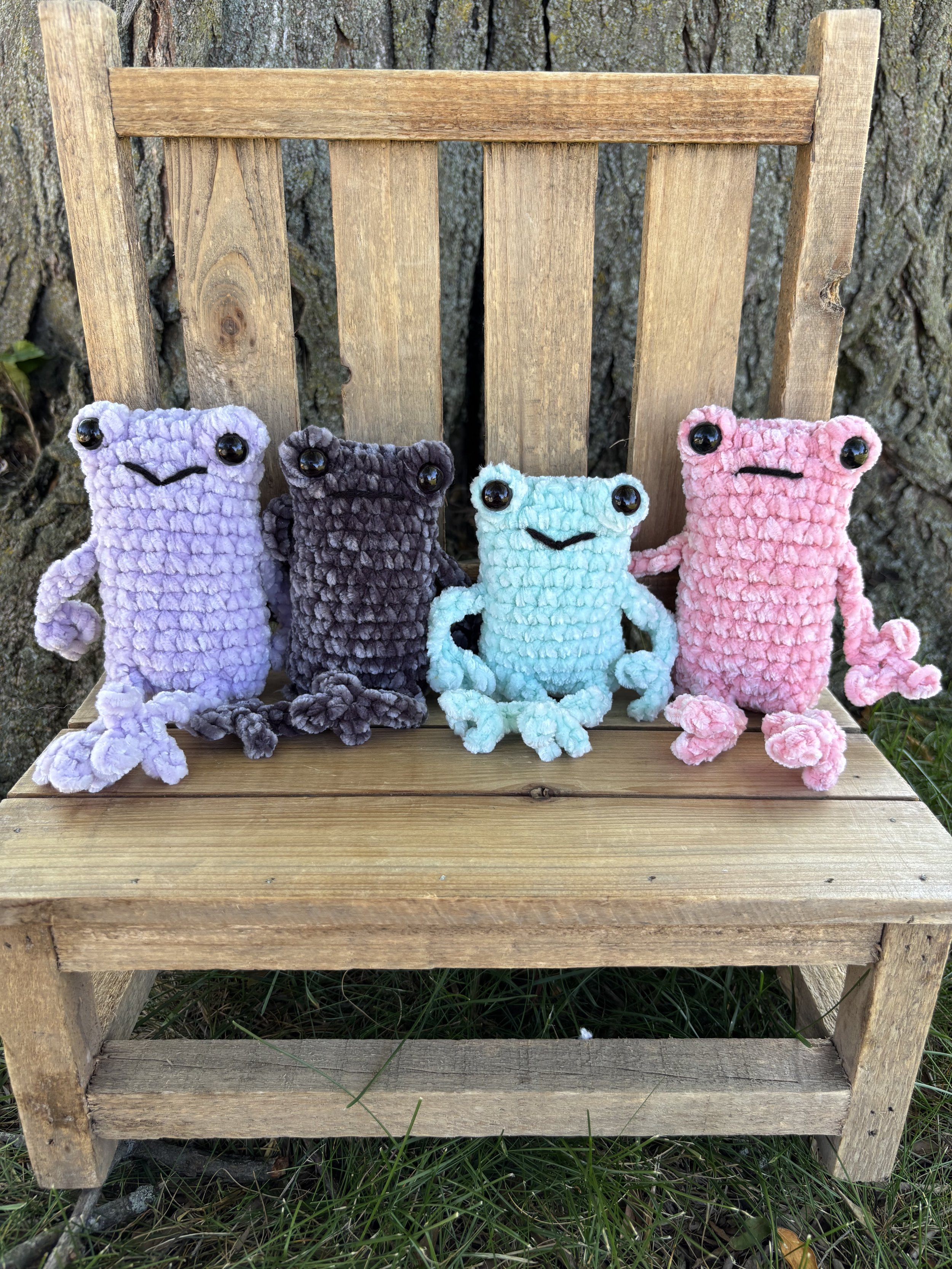 Four crochet velvet leggy frog plushies in lavender, grey, mint, and pink on wooden bench