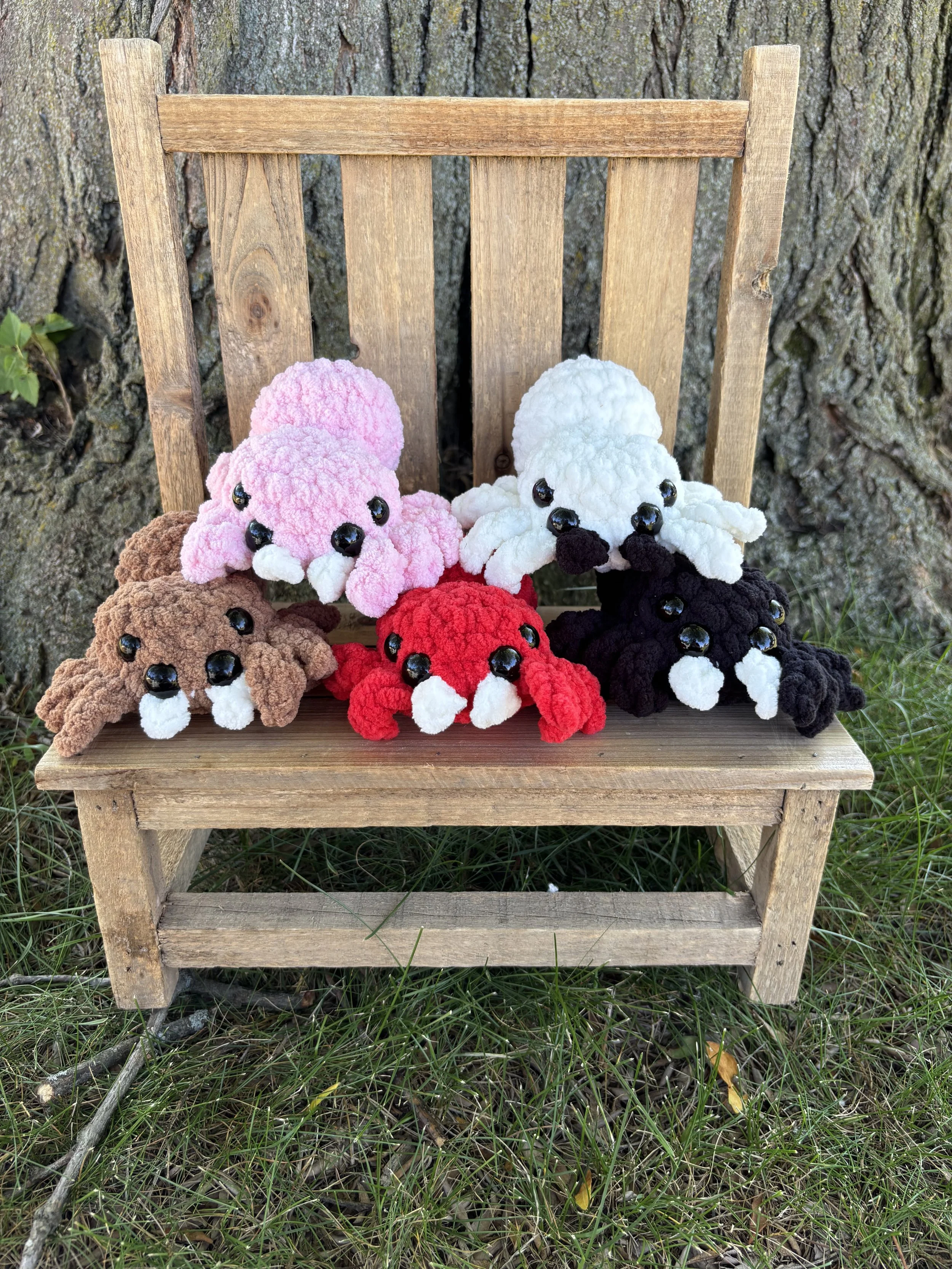 Five crochet jumping spider plushies in brown, black, red, pink, and white on wooden bench
