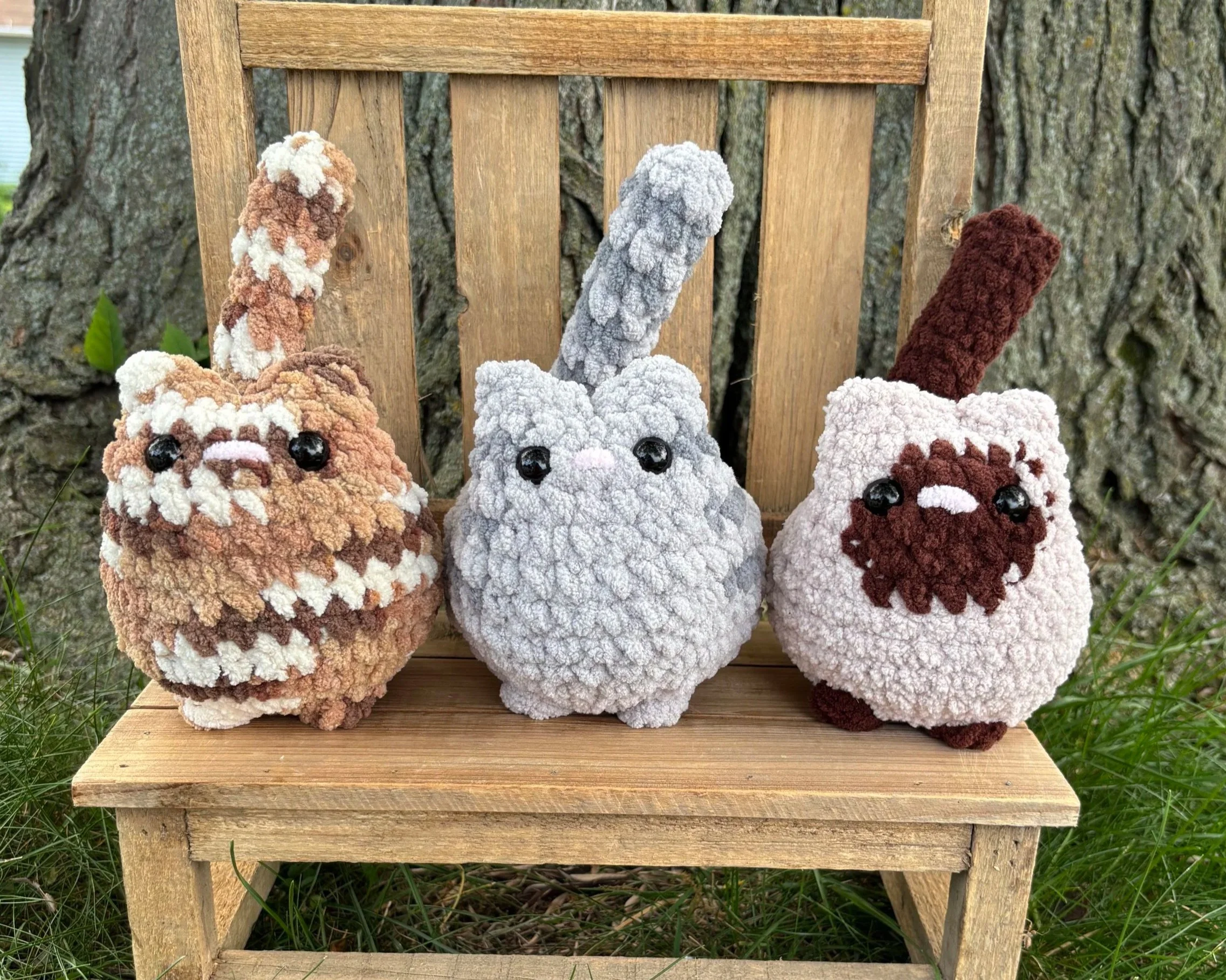 Crochet tabby and Siamese cat plushies sitting on wooden bench outdoors