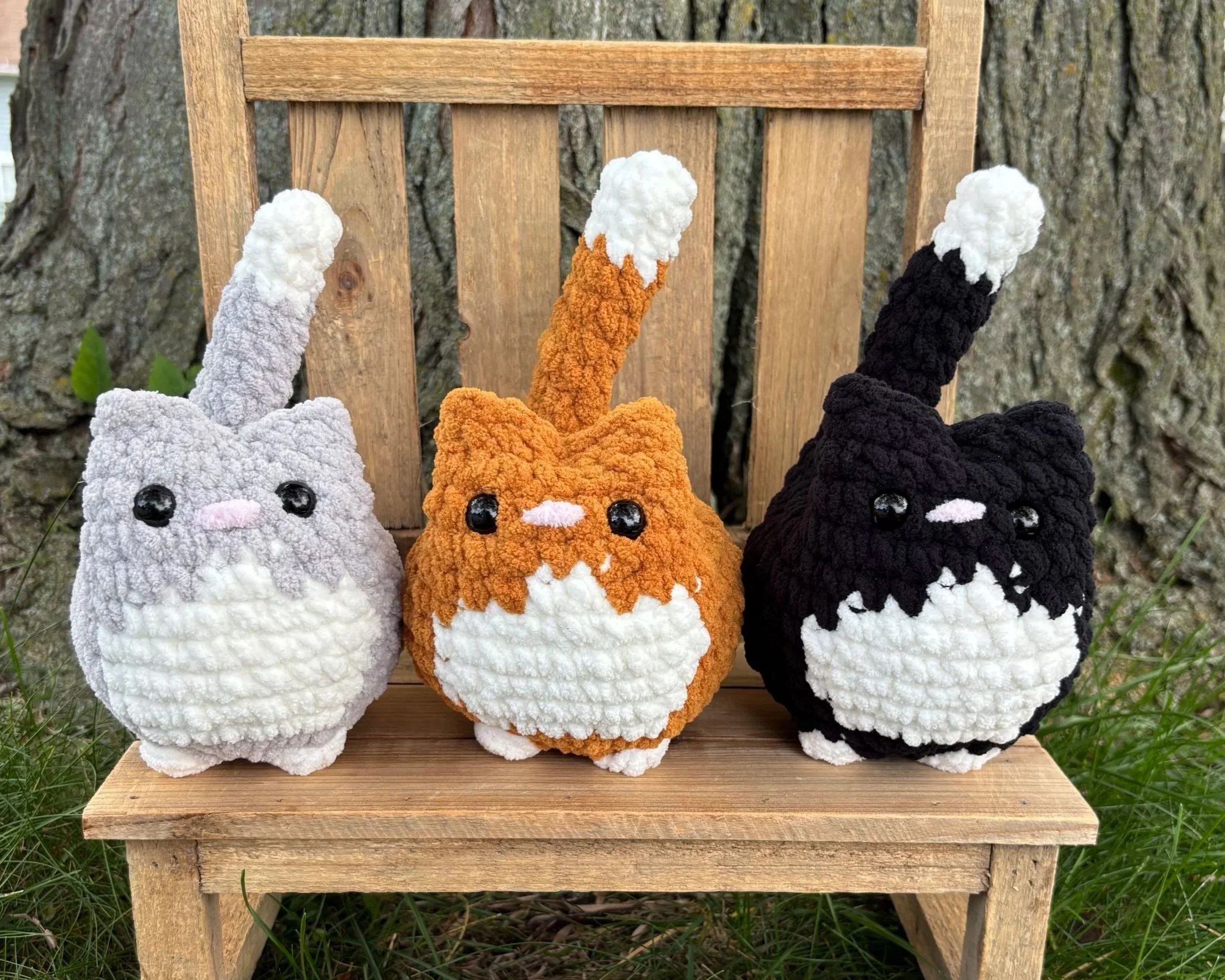 Crochet tuxedo cat plushies in grey, orange, and black on wooden bench