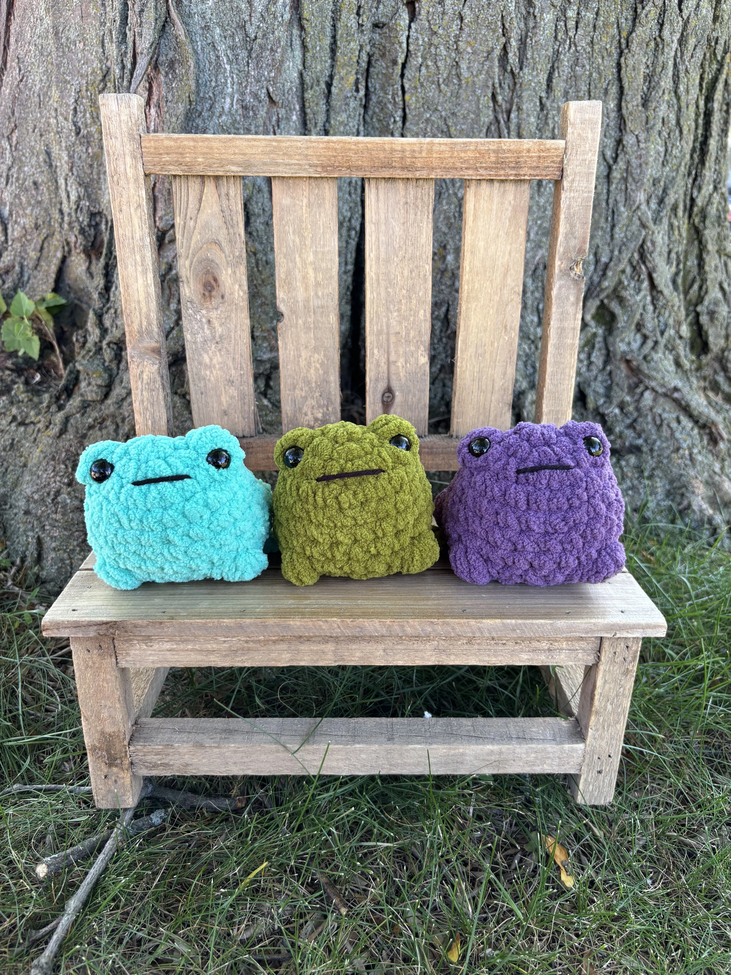 Three crochet jumping frog plushies in teal, olive green, and purple on wooden bench