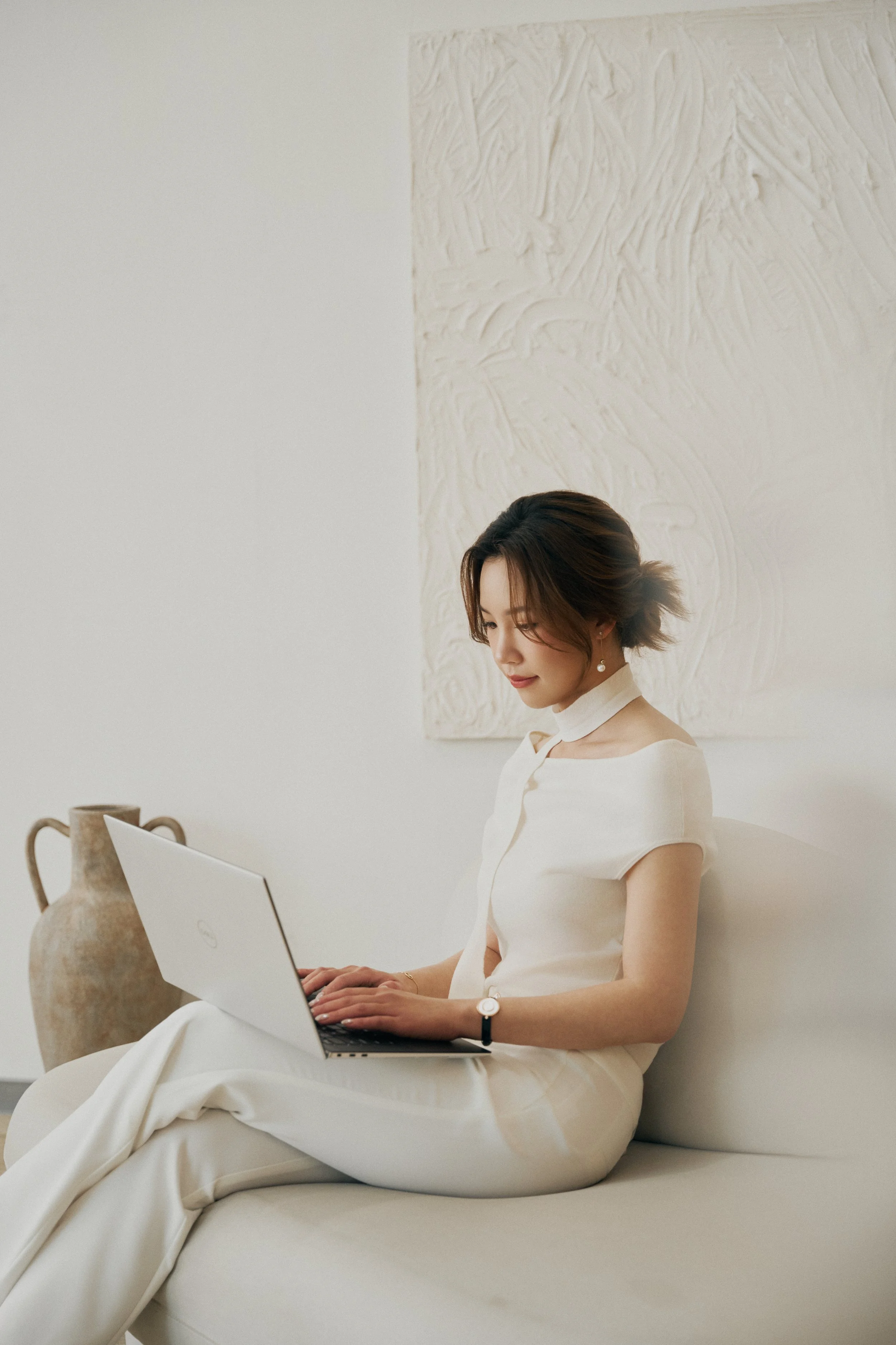 Interior designer wearing white outfit working on a laptop, seated on a light-colored couch, with a textured wall and vase in the background working on client work