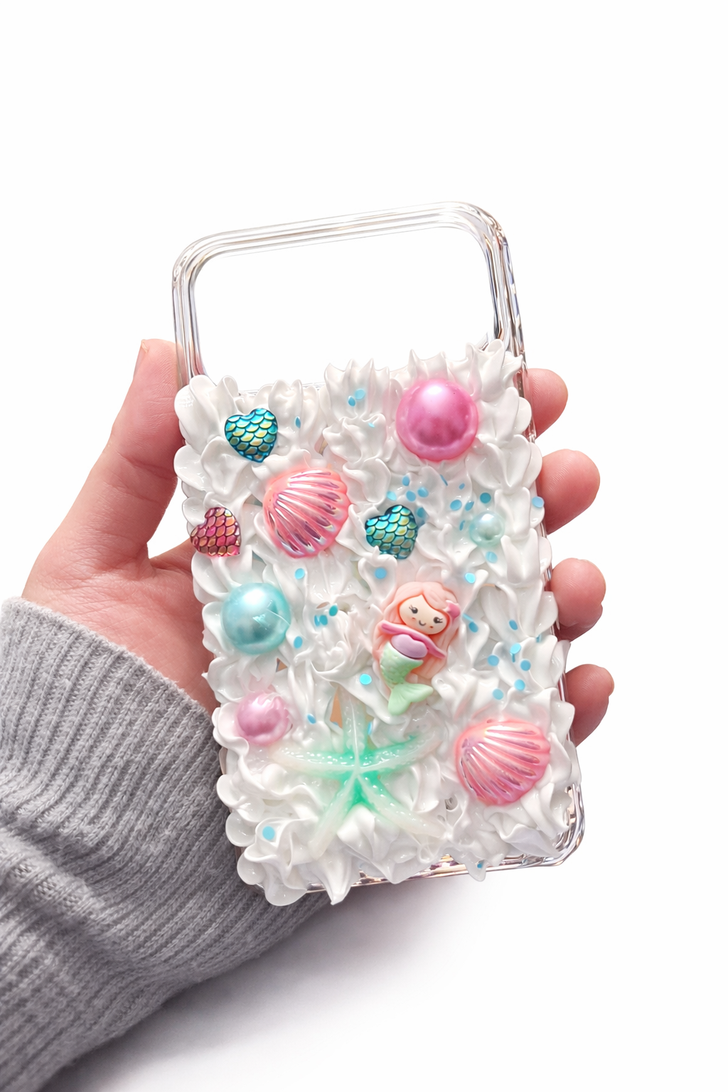 Decoden - Hand holding a phone case decorated with white whipped texture, pink, blue, and green seashells, a mermaid figure, and star-shaped decorations, all on a white background.