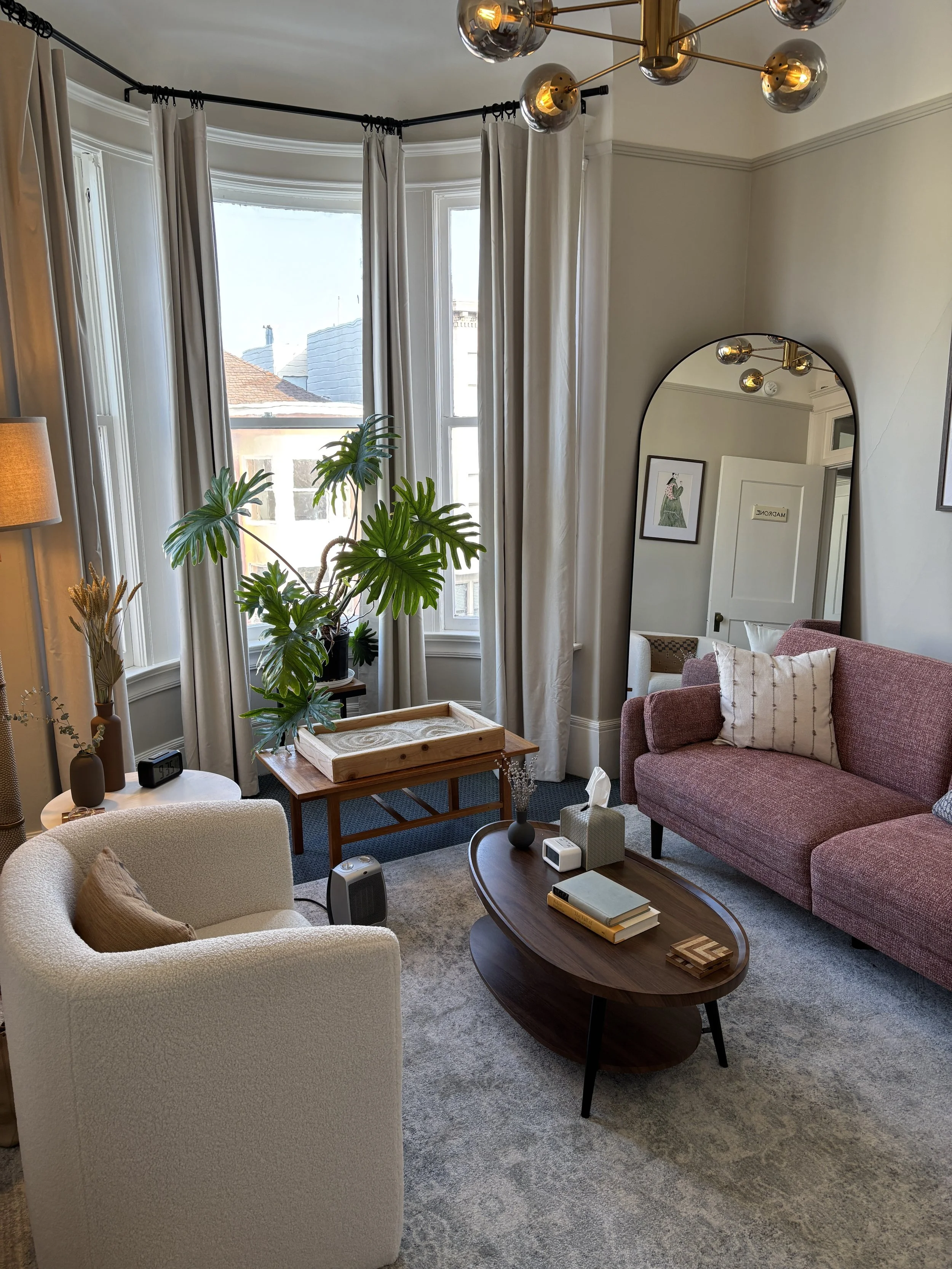 Therapy room with red sofa, brown coffee table, white therapist chair, mirror, large windows with floor length tan curtains