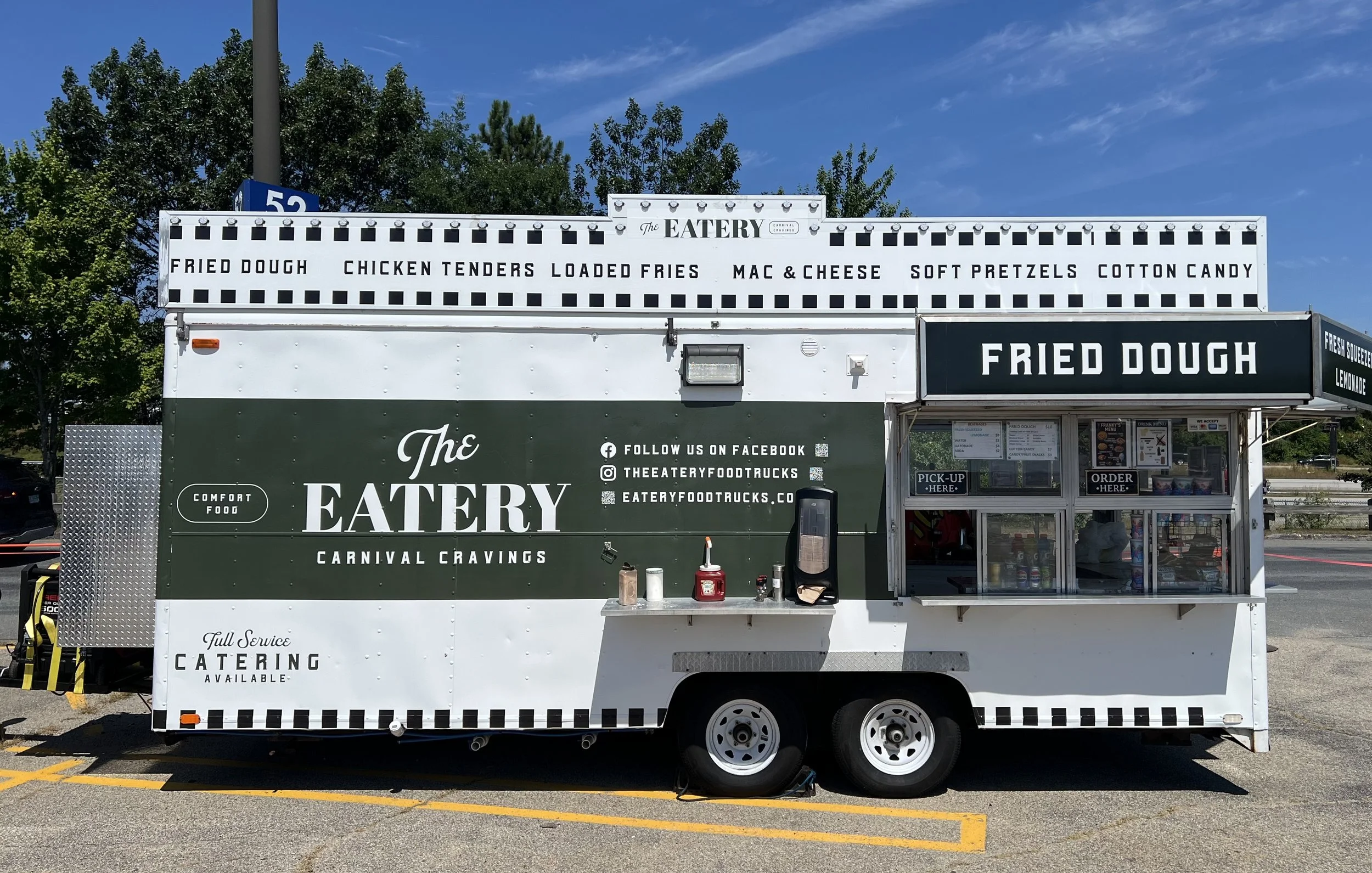 A food concession trailer with signs advertising tenders, fries, cotton candy, and other snacks, parked near a tennis court with a fence, under an overcast sky.