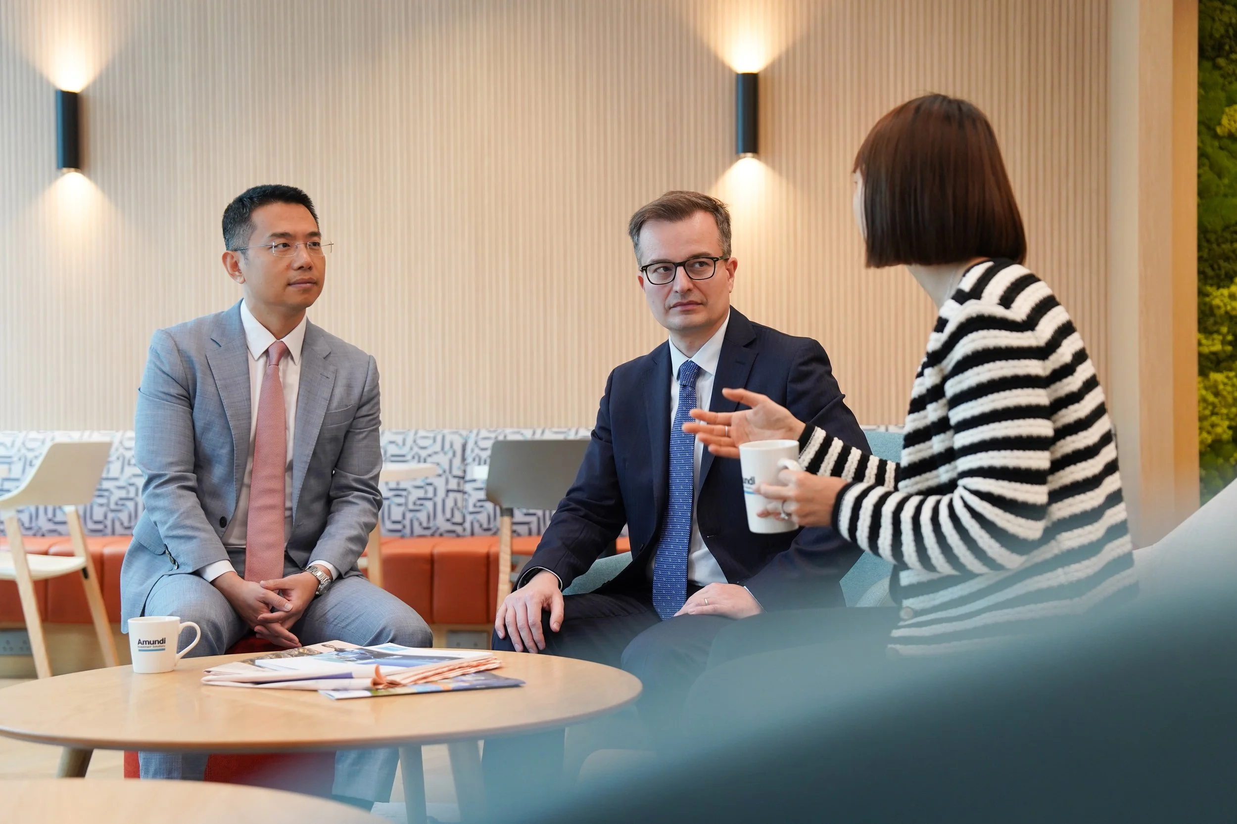 Three business professionals having a discussion in a modern office lounge, two men in suits talking to a woman in a striped sweater holding a coffee mug.
