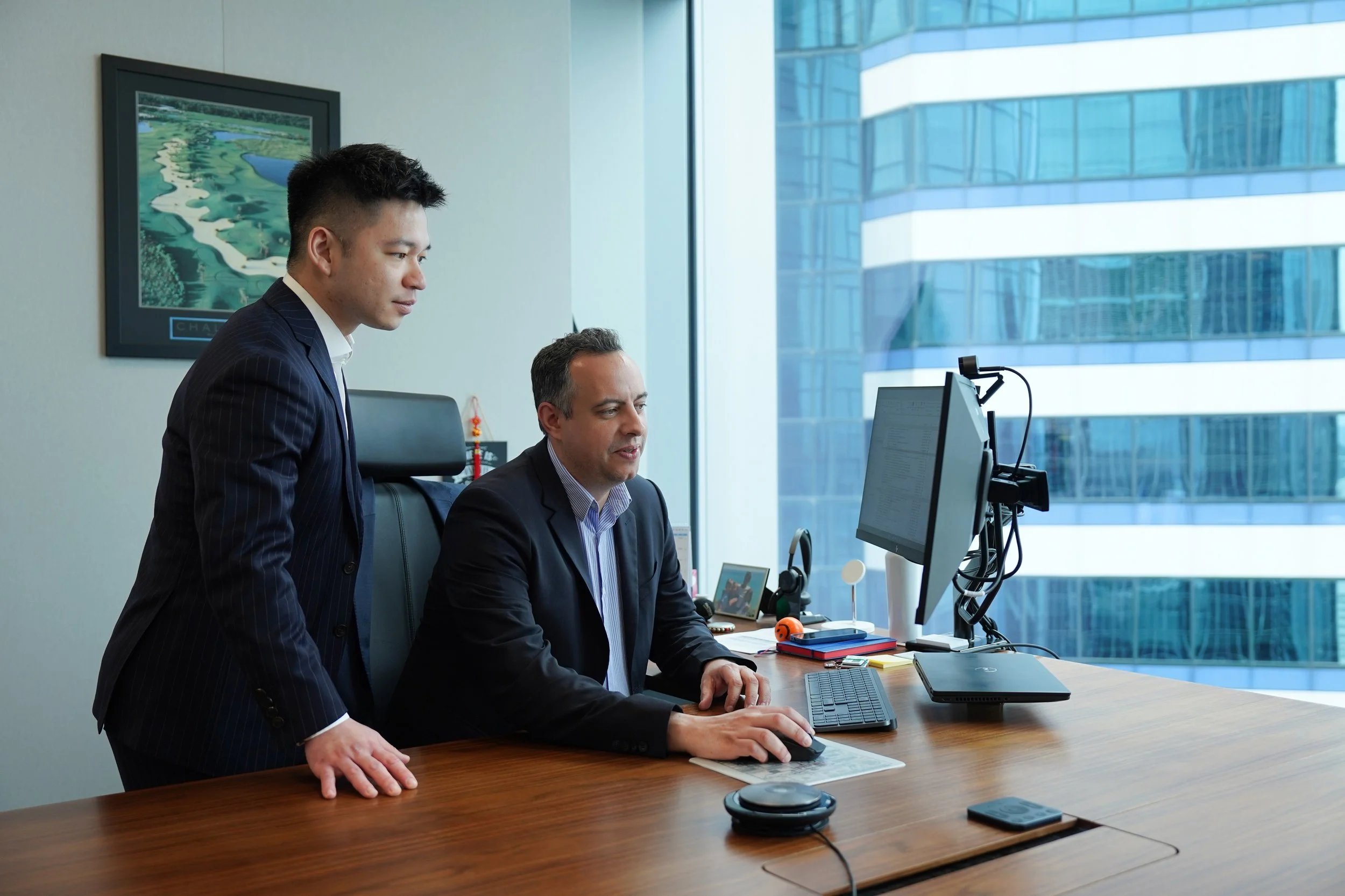 Two businessmen in suits working together in a modern office, one sitting at a desk using a computer and the other standing beside him, looking at the screen, with a view of a city skyscraper outside the window.