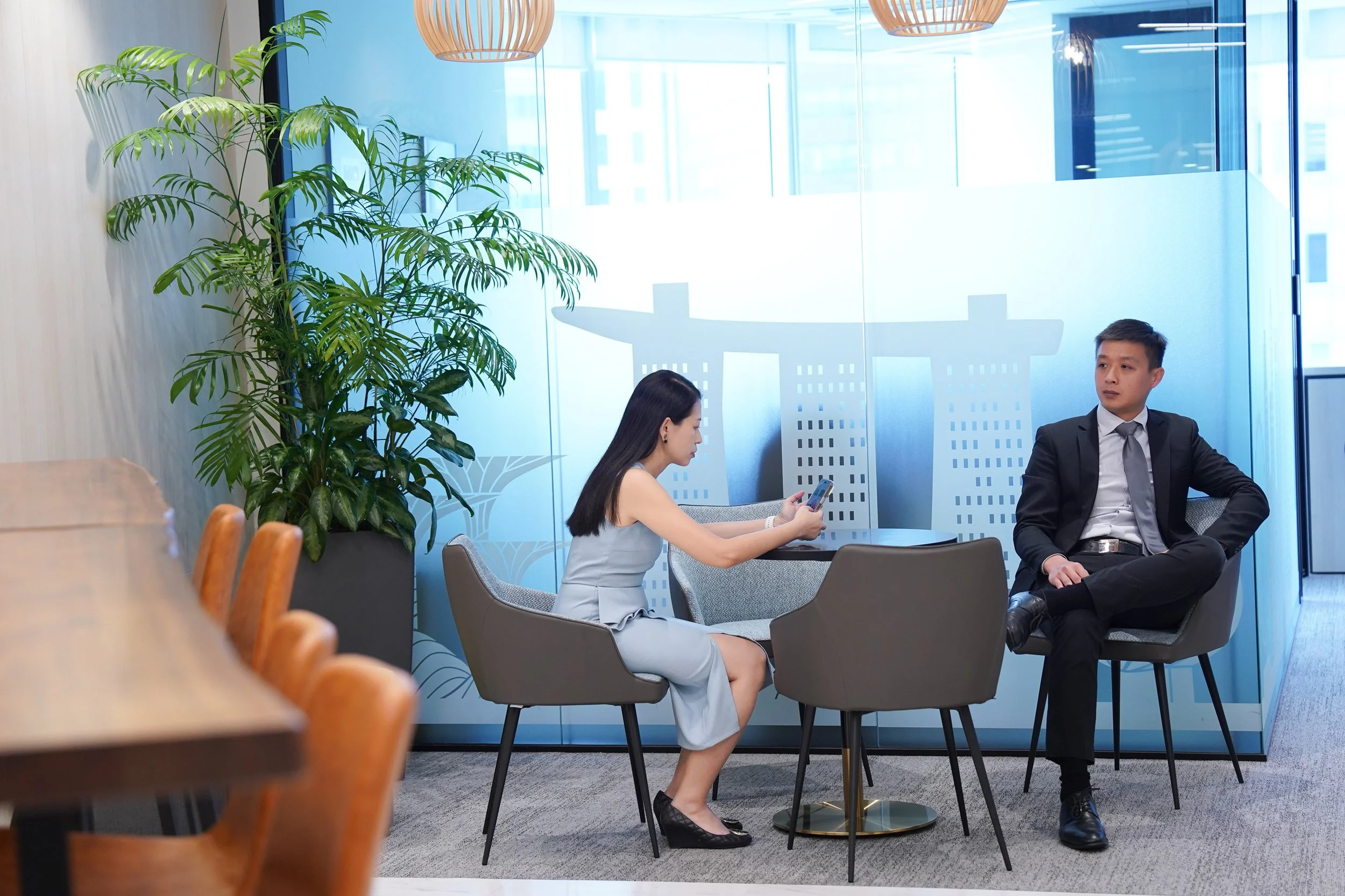 A woman in a light gray dress is sitting at a small table, looking at her phone, while a man in a black suit with a gray tie and white shirt, is seated with his arm resting on the chair back, in a modern office space with glass walls and a large gree