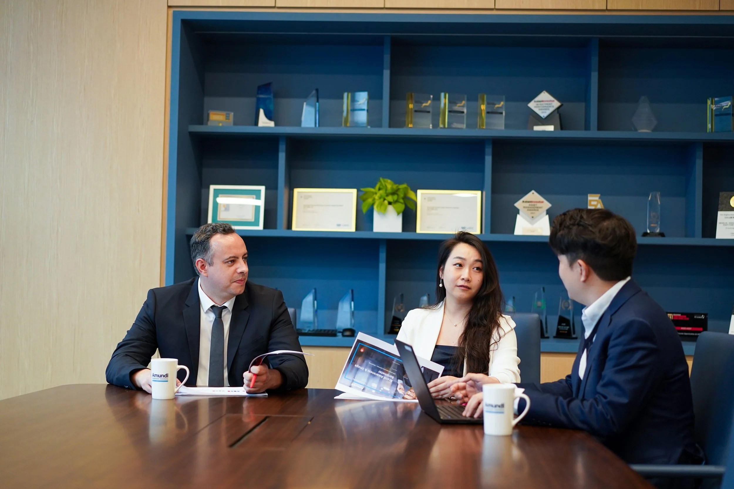 Three business professionals sitting at a conference table, having a discussion, with awards and certificates displayed on a blue wall behind them.