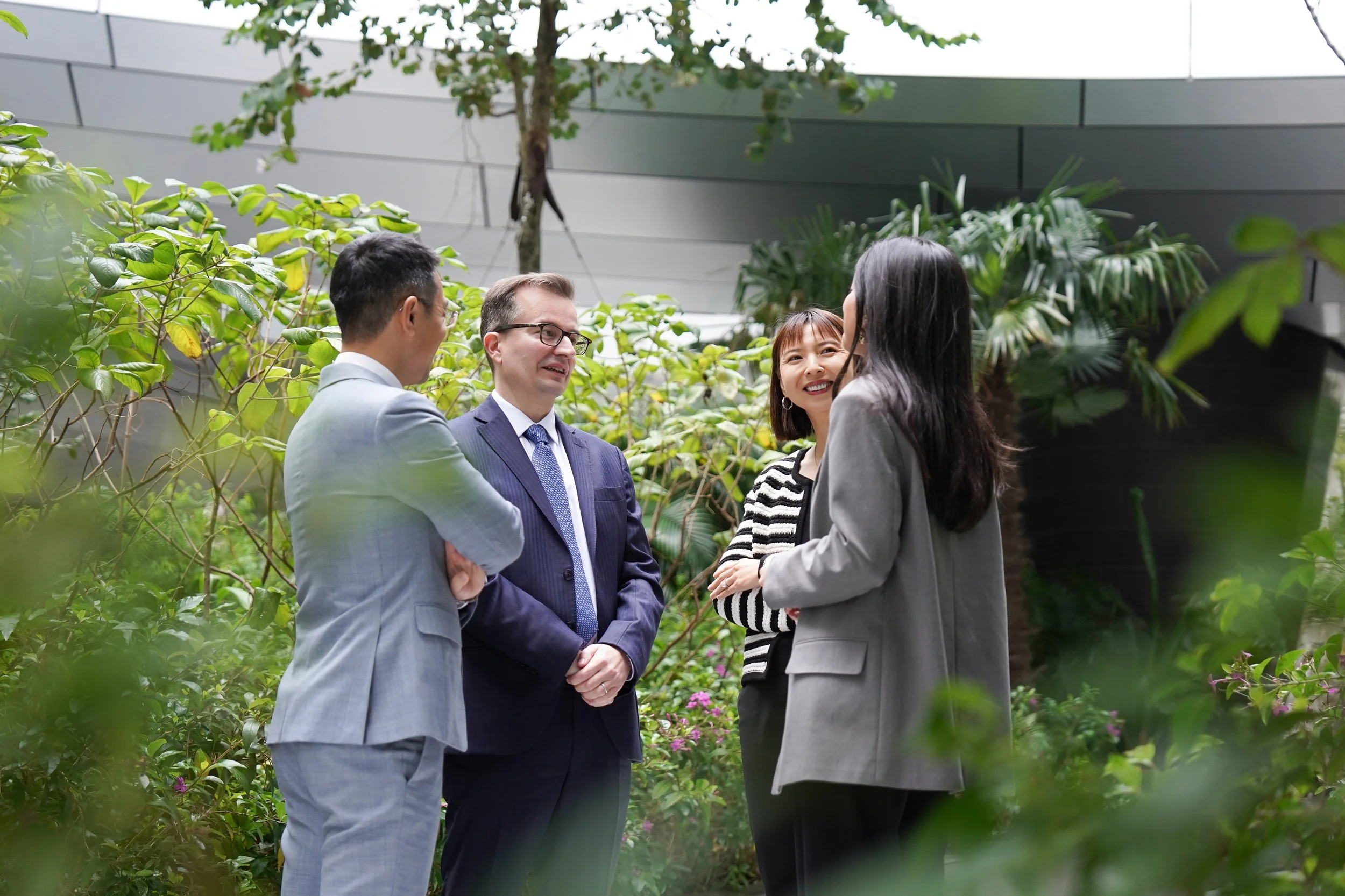 Four businesspeople, two men and two women, are having a conversation outdoors among greenery and plants, dressed in professional attire.
