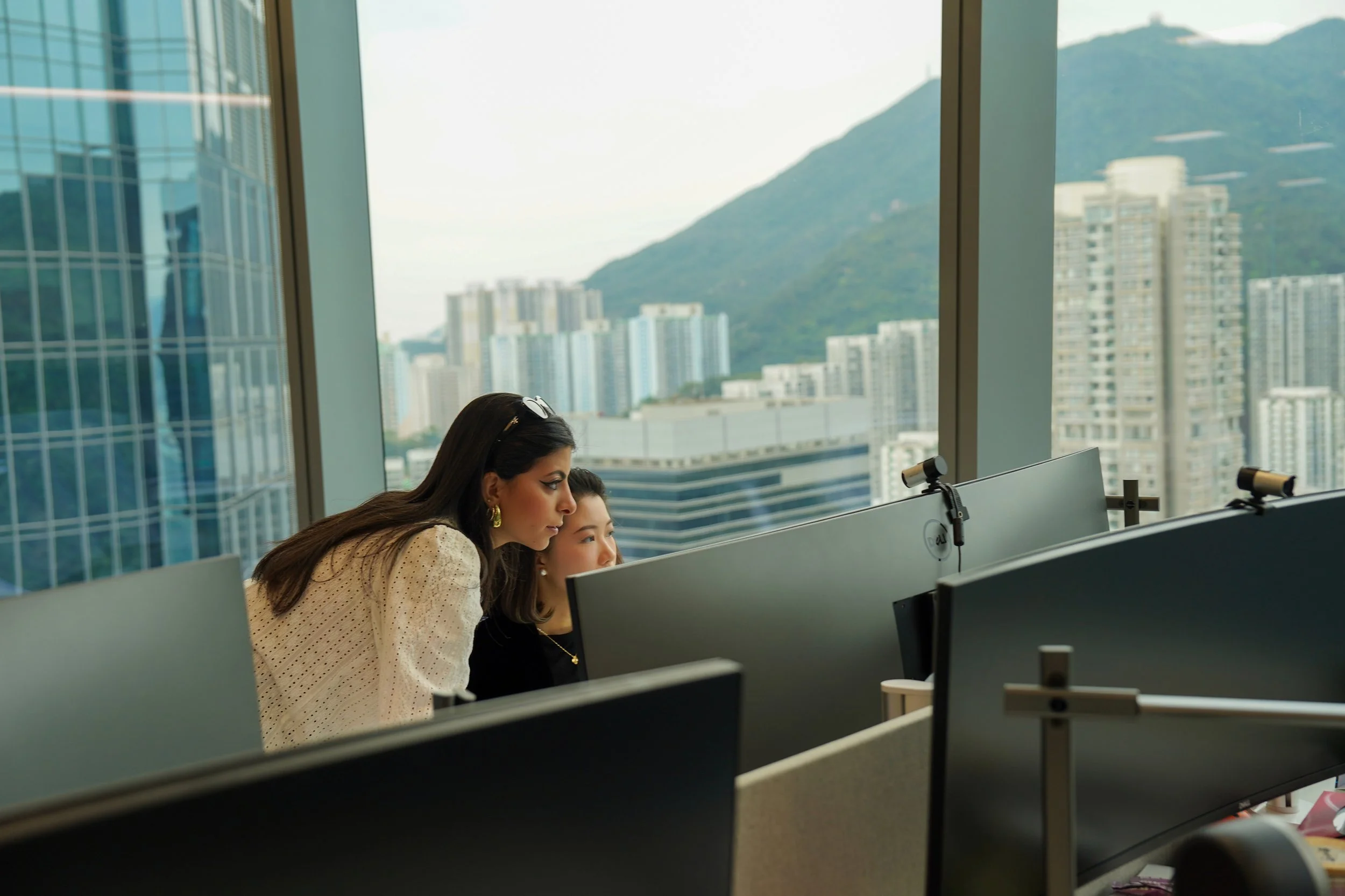 Two women work at computer stations in an office with large windows showing a city skyline and mountains in the background.