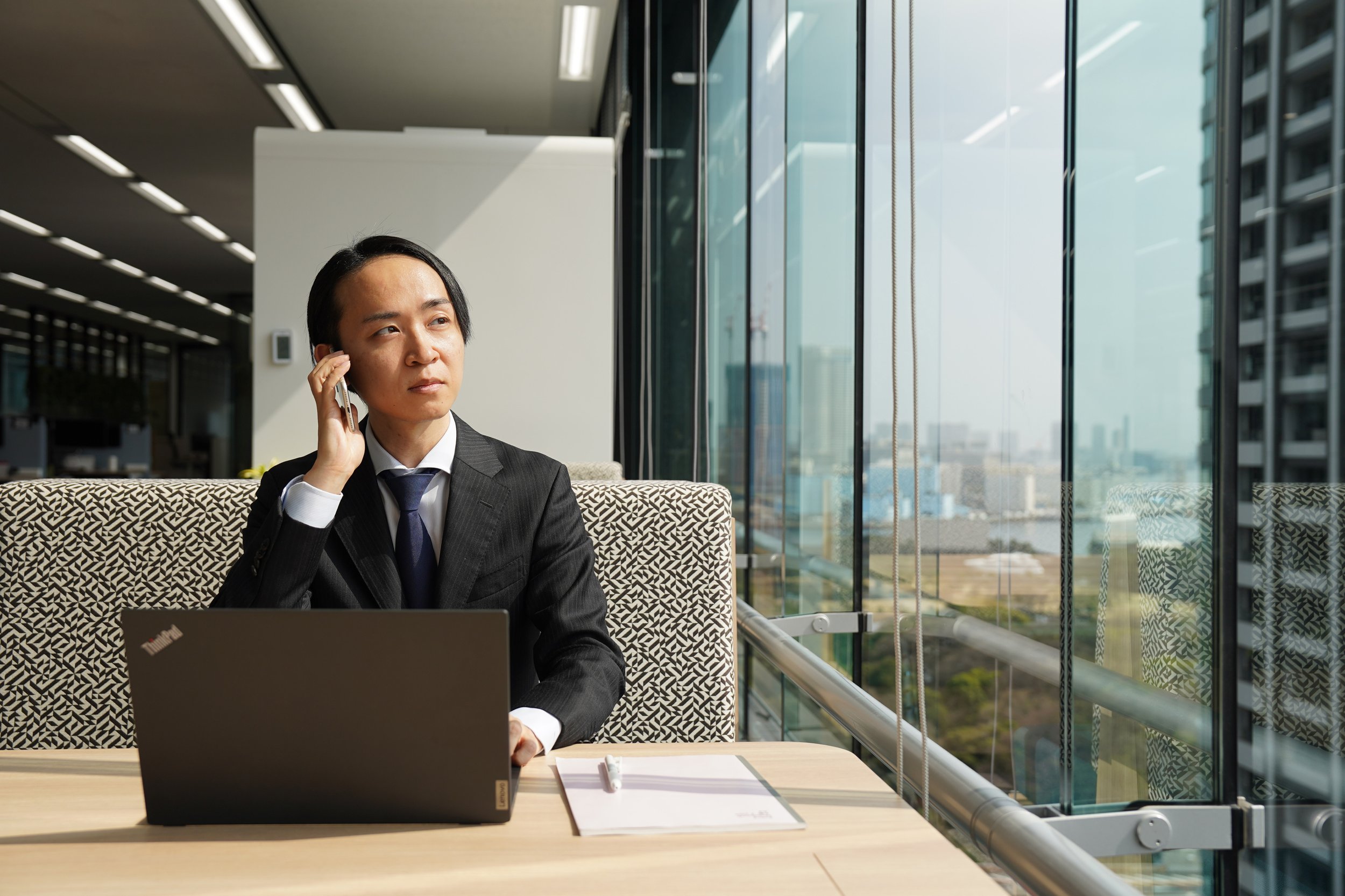 Businesswoman in suit sitting at a desk using a laptop and talking on a cell phone in a modern office with large glass windows overlooking a cityscape.