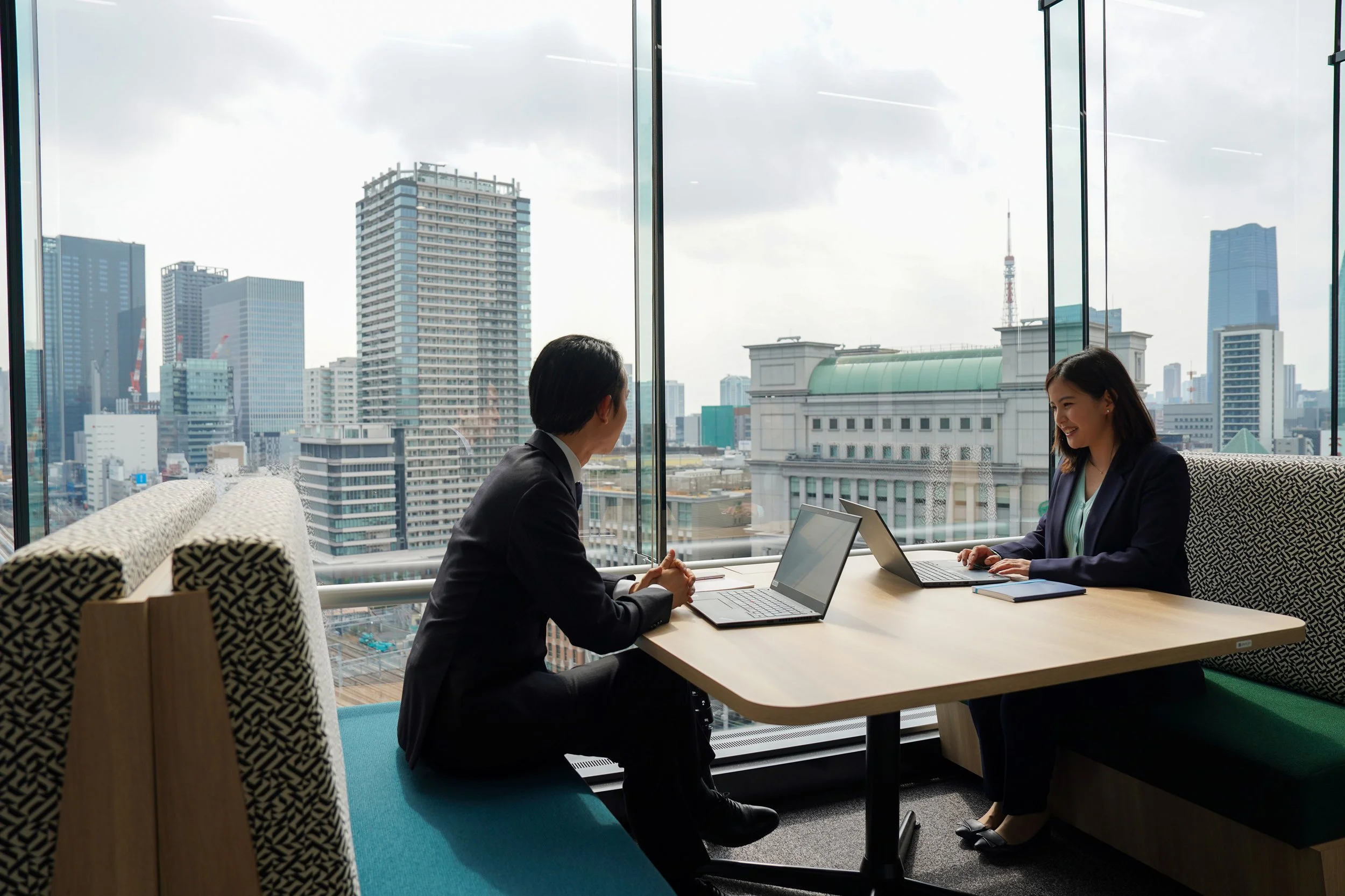 A man and a woman having a business meeting in a modern office with floor-to-ceiling windows showing a cityscape of tall buildings in the background.