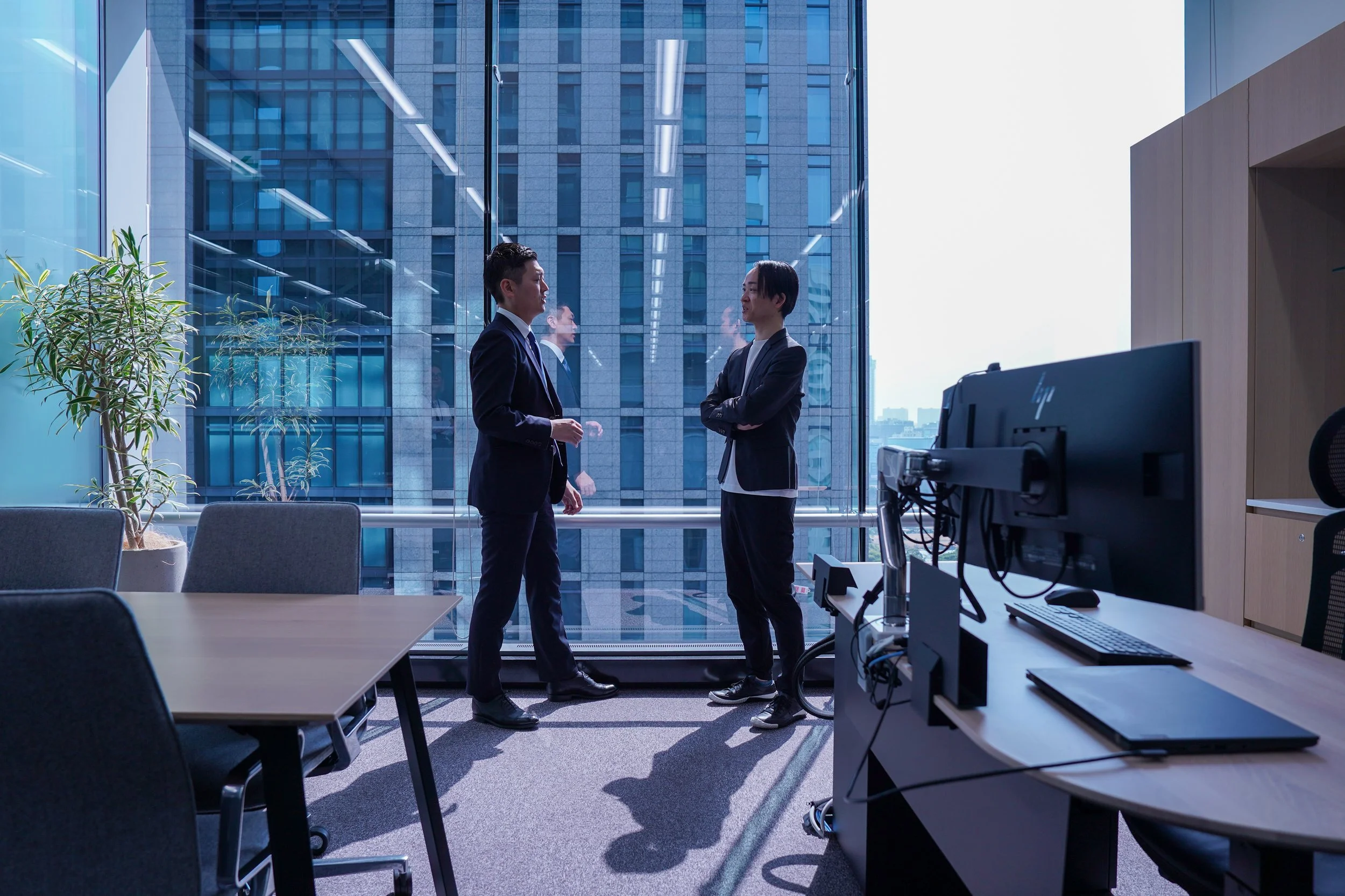 Two men in business suits standing and talking in a modern office with a large window overlooking city buildings.