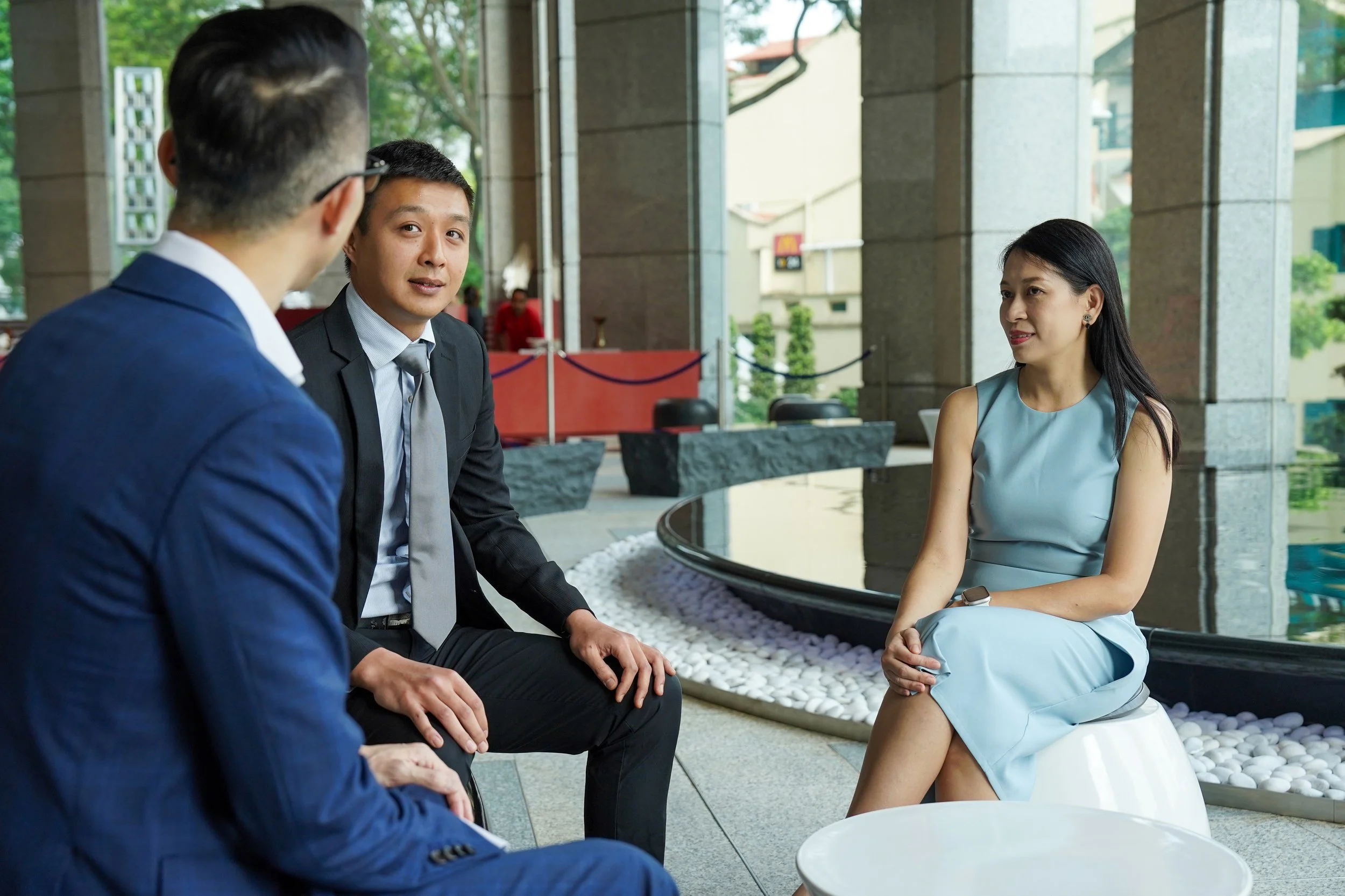 Three people having a conversation in a modern lobby near a water feature, with two men in suits and a woman in a light blue dress.