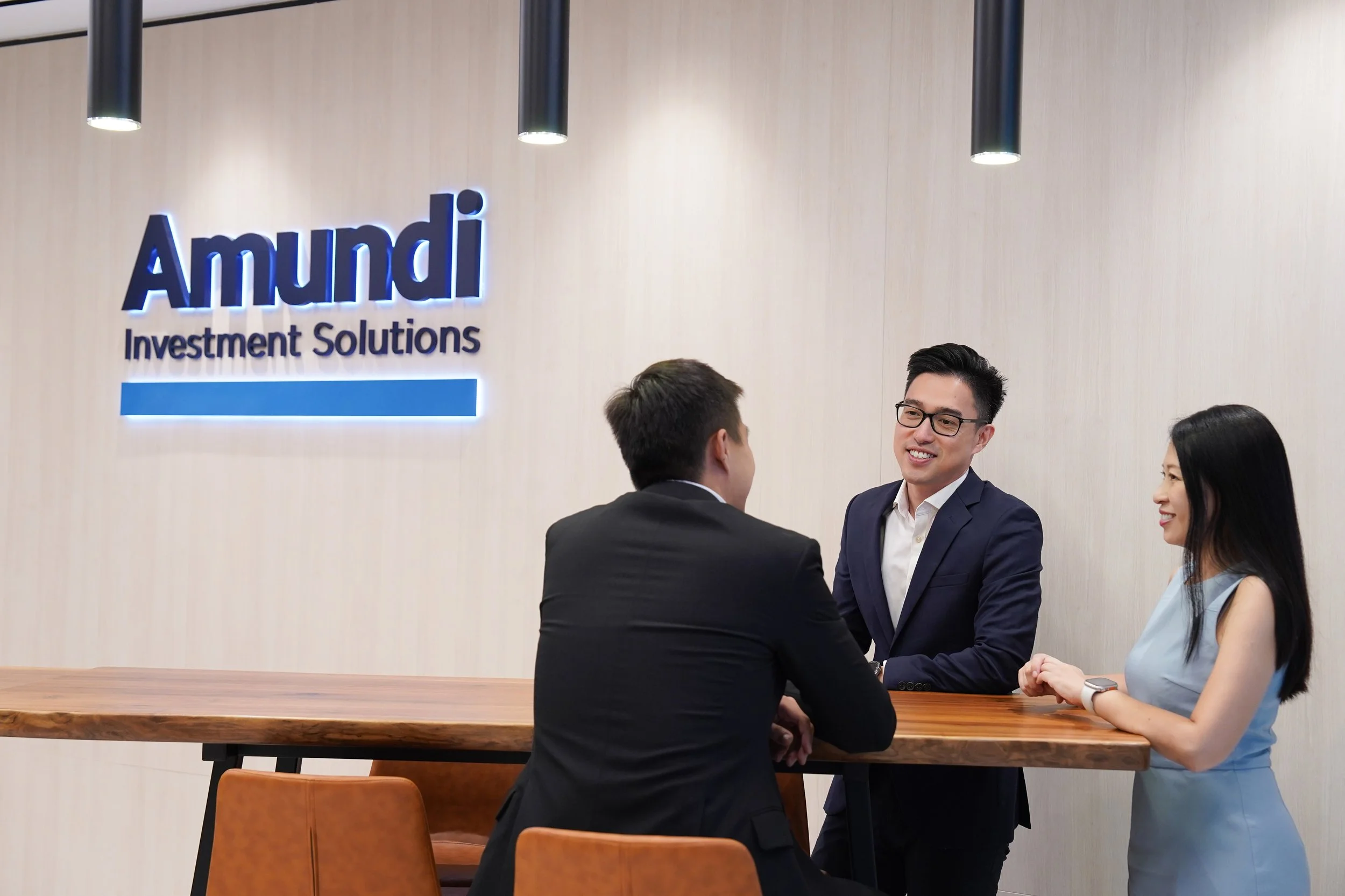 Three business professionals having a discussion at a high wooden table in an office with a sign on the wall that reads "Amundi Investment Solutions." The man on the left is wearing a black suit, the man in the middle is wearing glasses and a dark bl