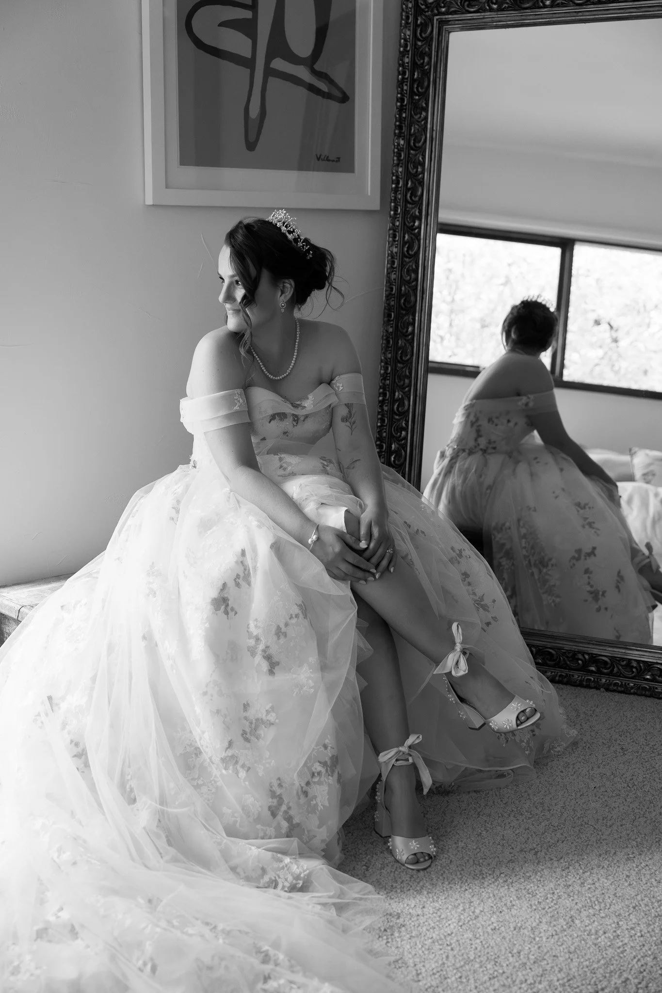A bride in a wedding dress sitting near a large mirror, with her reflection showing from the back, in a softly lit room.