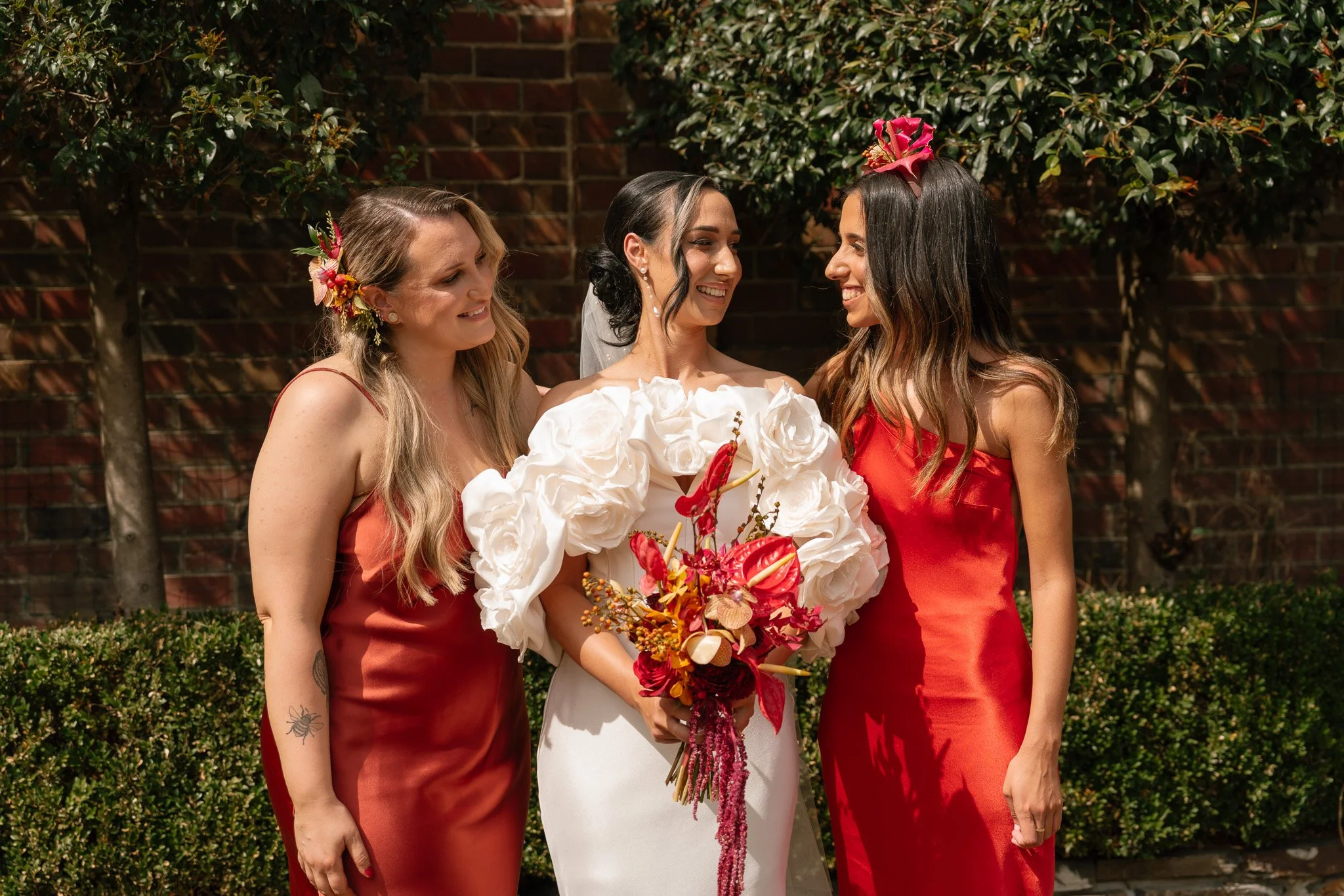 Three women at a wedding, one in a white dress holding a bouquet of red, pink, and white flowers, with two women in red dresses standing beside her. They are outdoors with bushes and a brick wall in the background, smiling and looking at each other.