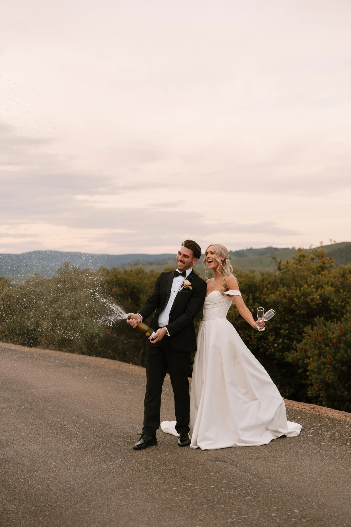 A newlywed couple dressed in wedding attire celebrating outdoors with champagne, spraying it and laughing.