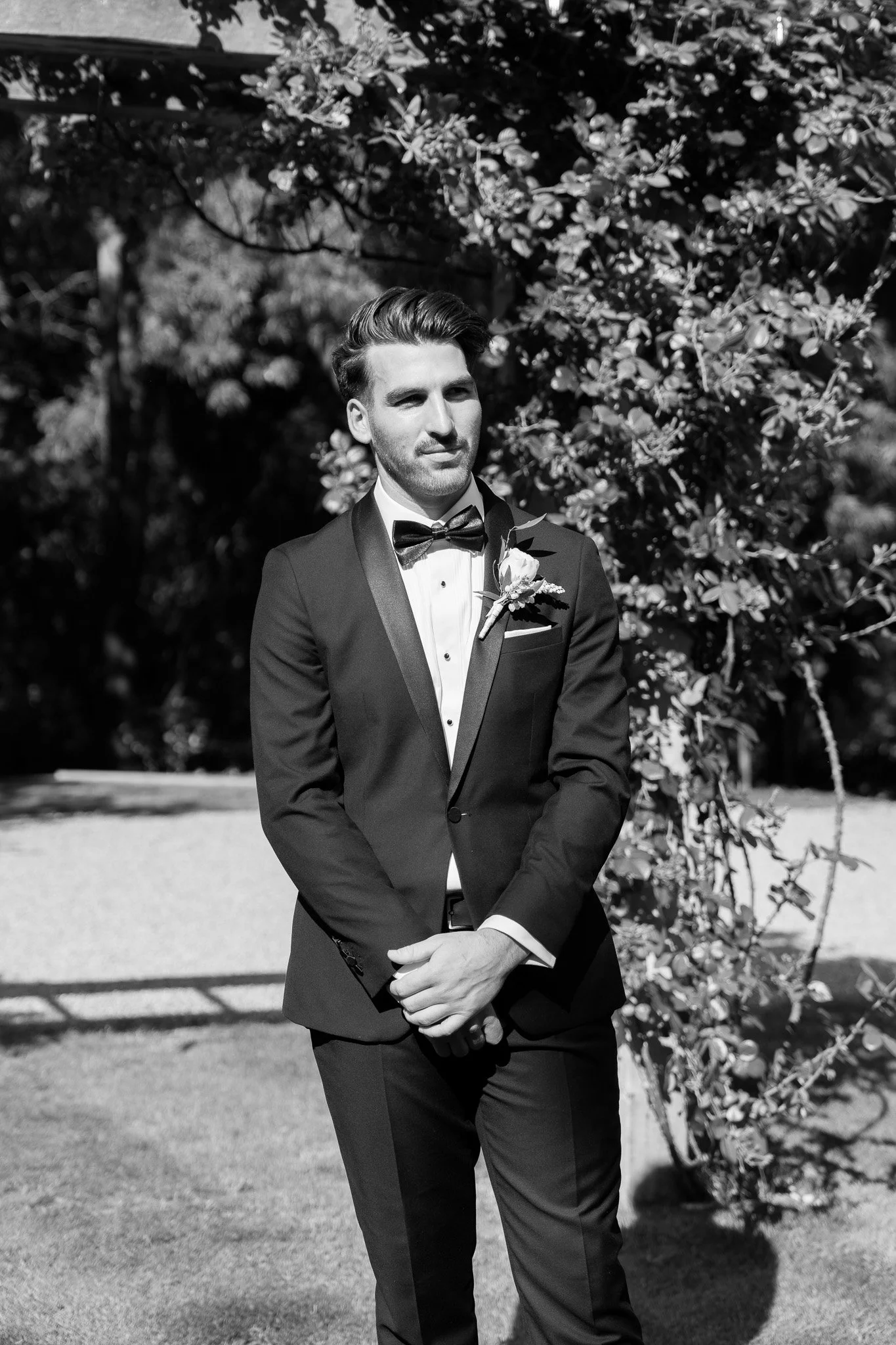 A man dressed in a black tuxedo with a bow tie, standing outdoors in front of trees, looking to the side, in a black and white photograph.