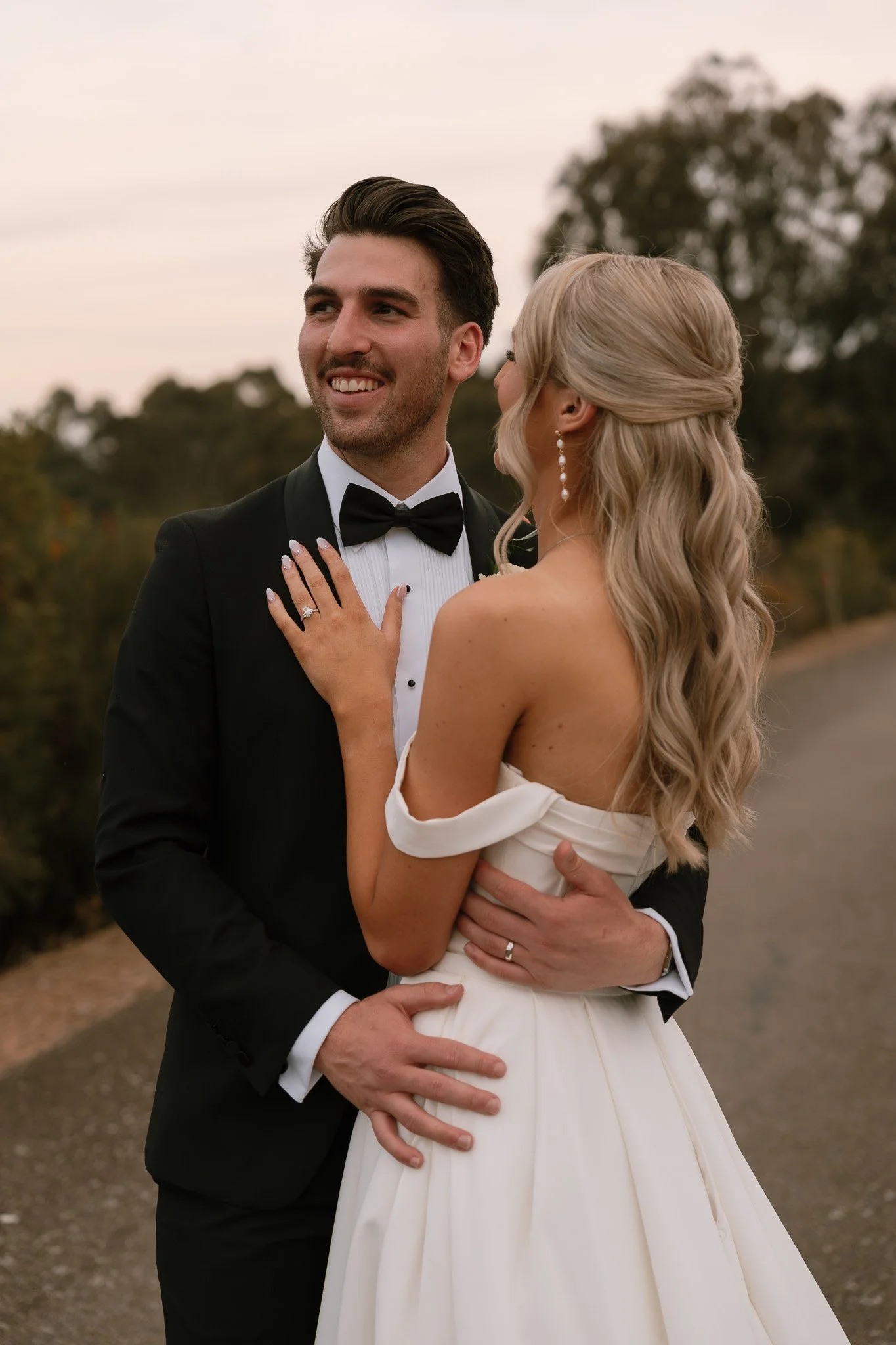 A newlywed couple dressed in wedding attire standing outdoors on a country road, embracing and gazing at each other during daylight.