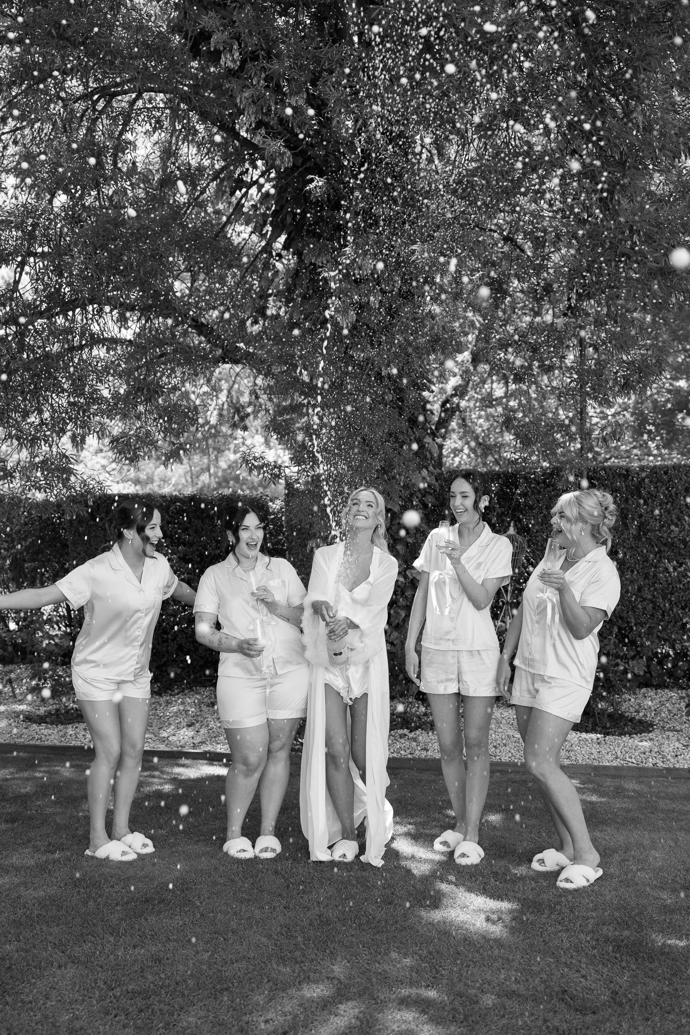 A group of five women celebrating outdoors, wearing matching pajamas, popping champagne under a large tree.
