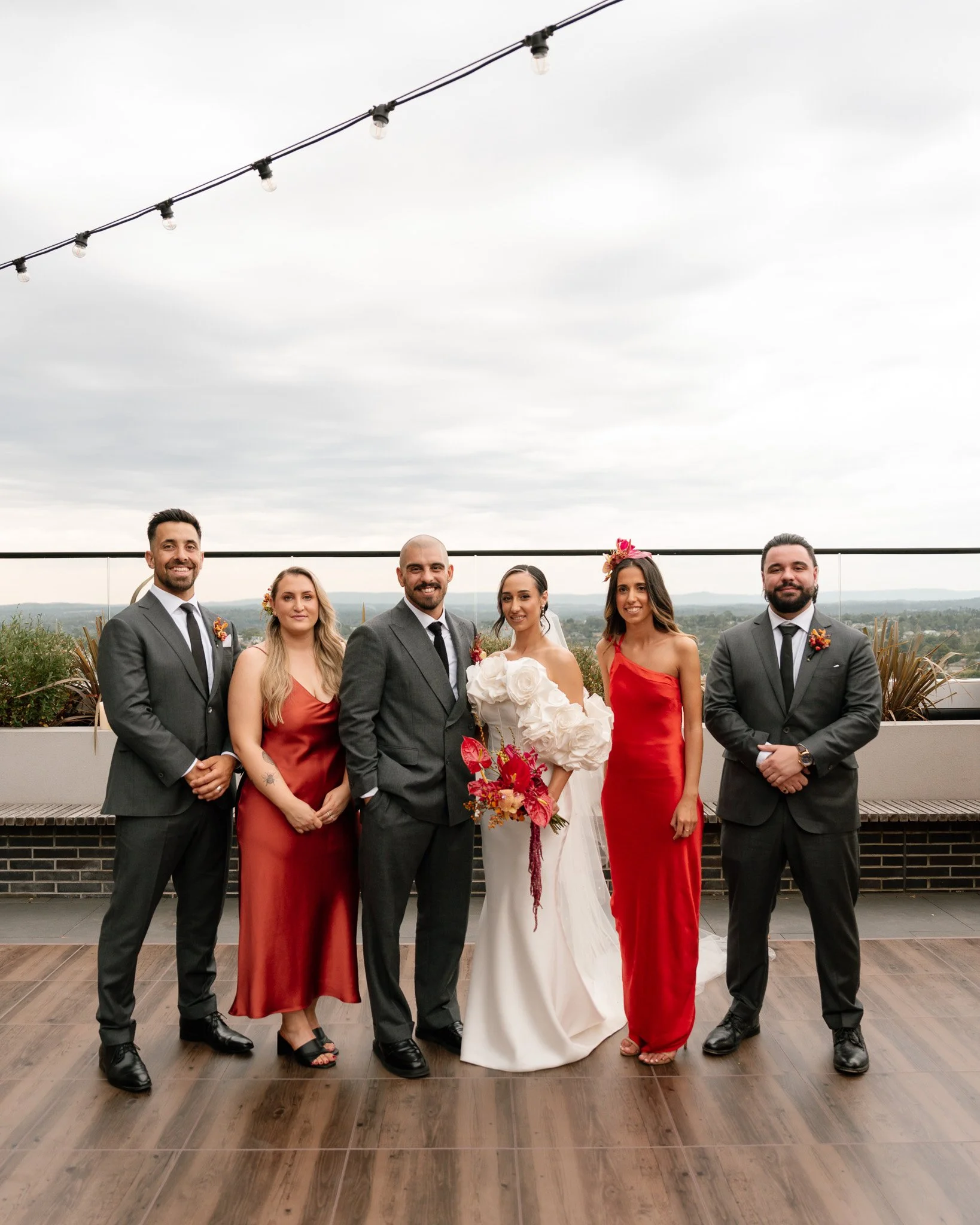 Wedding party consisting of a bride in a white dress holding a bouquet, three men in gray suits with ties, and two women in red dresses, standing outdoors on a rooftop with cityscape in the background and an overcast sky.