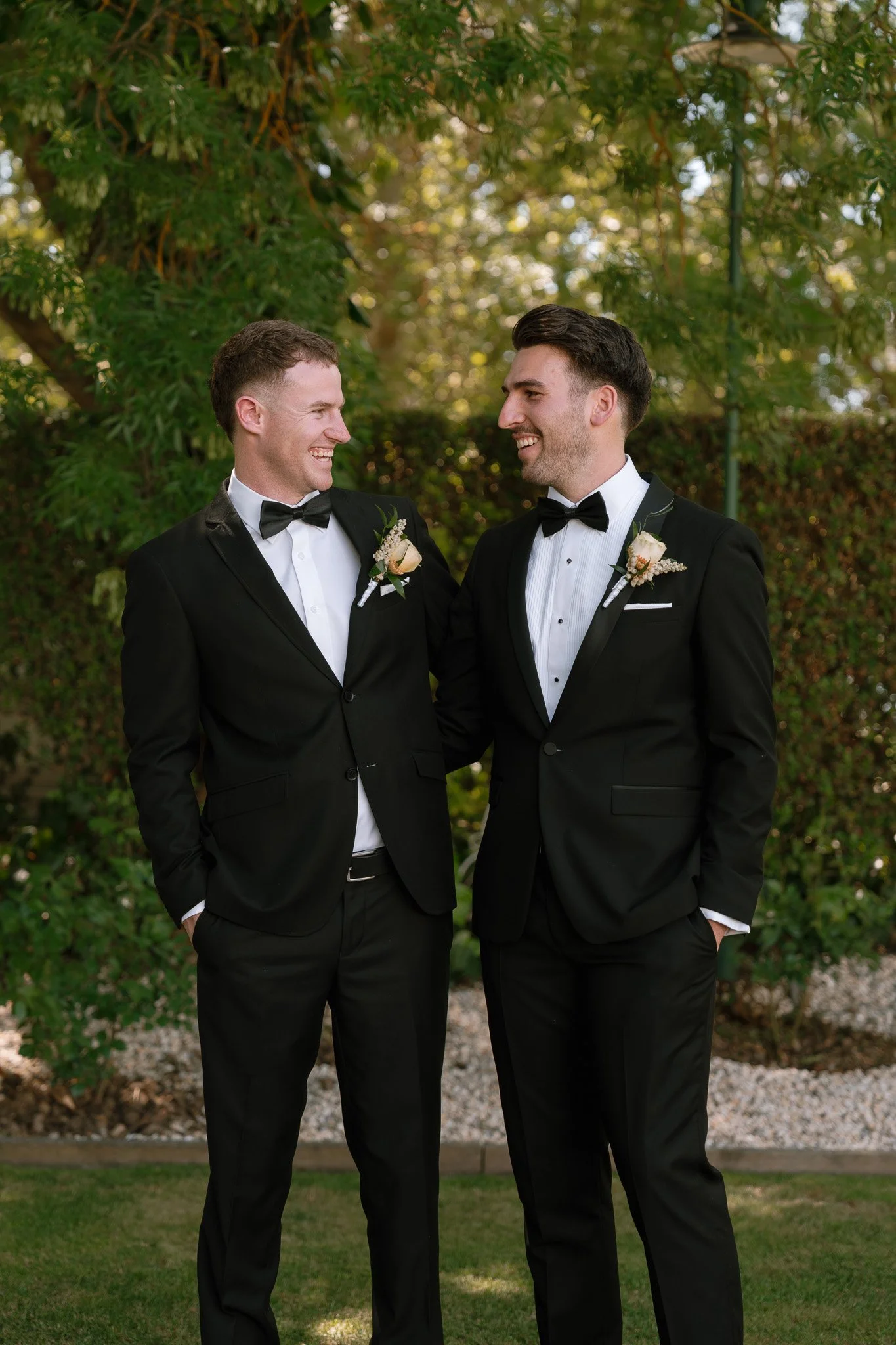 Two men in black tuxedos with bow ties, boutonnières, smiling and standing outdoors with greenery in the background.